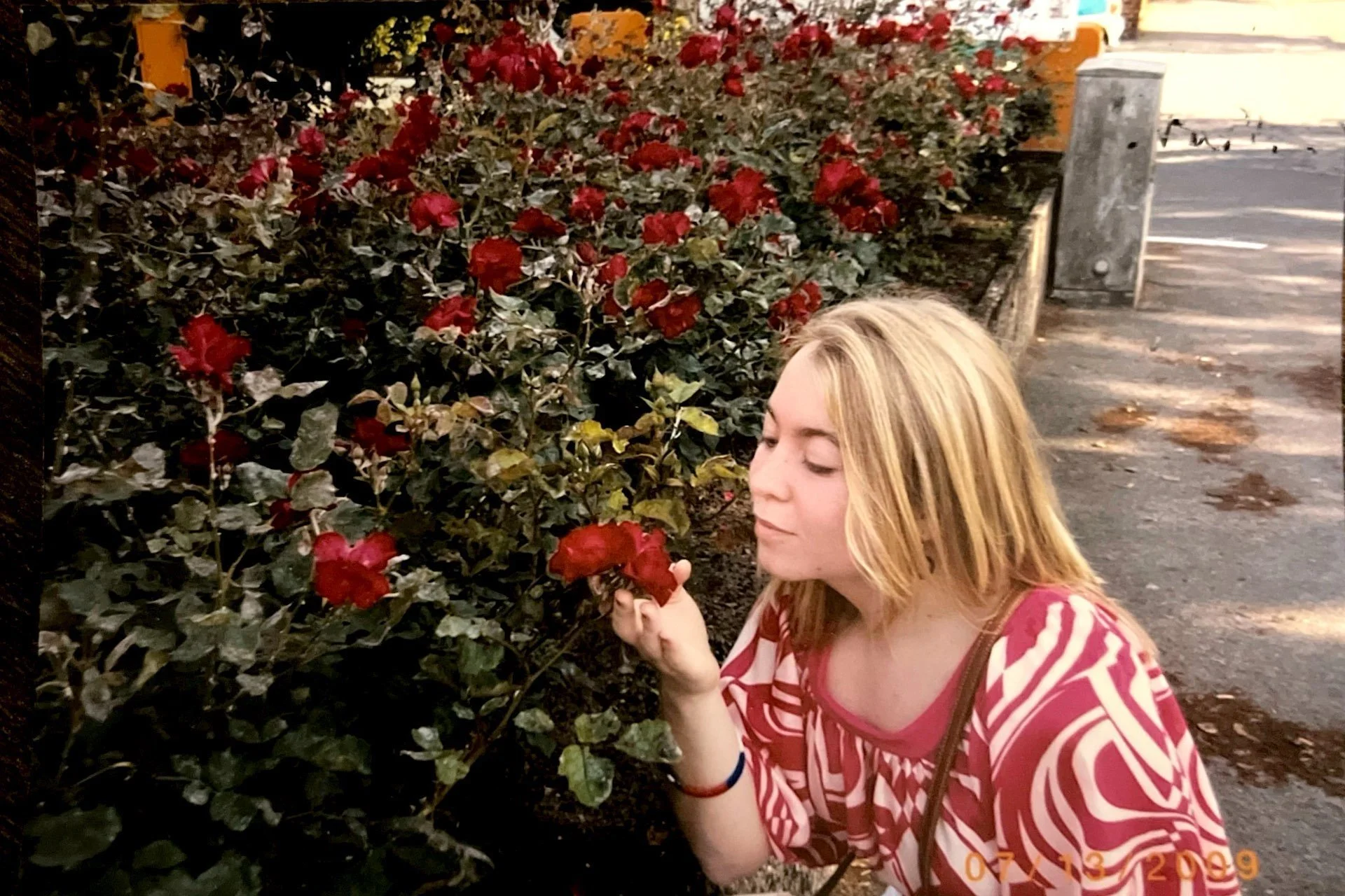 A young woman with blonde hair, wearing a red and white striped shirt, smells a red flower next to a bush with many red flowers, in an outdoor setting near a parking lot and a sidewalk.