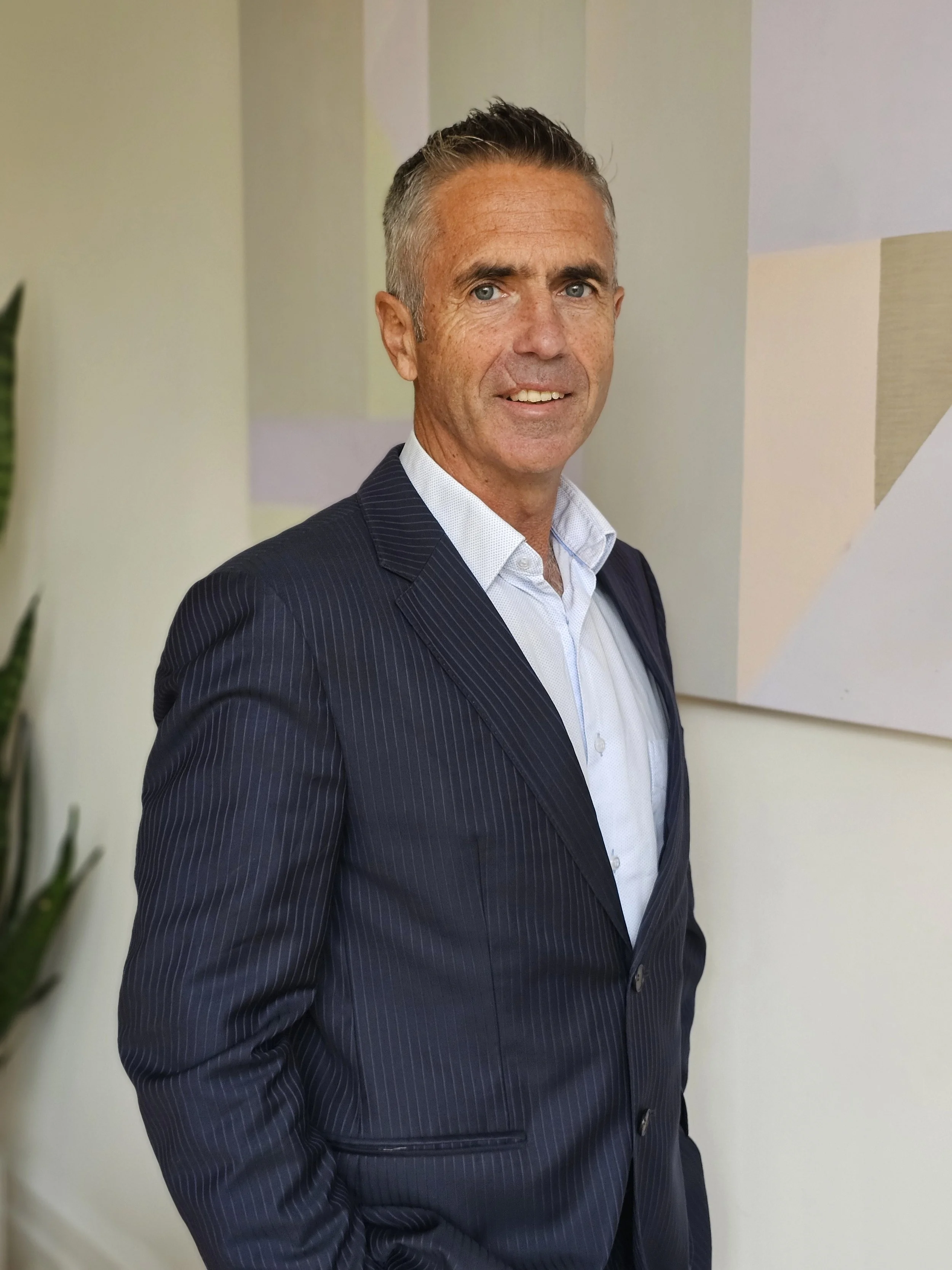 Mid-aged man with short gray hair, light skin, wearing a dark pinstripe suit and white shirt is smiling while standing indoors near a wall with abstract art. There is a green plant partially visible on the left.