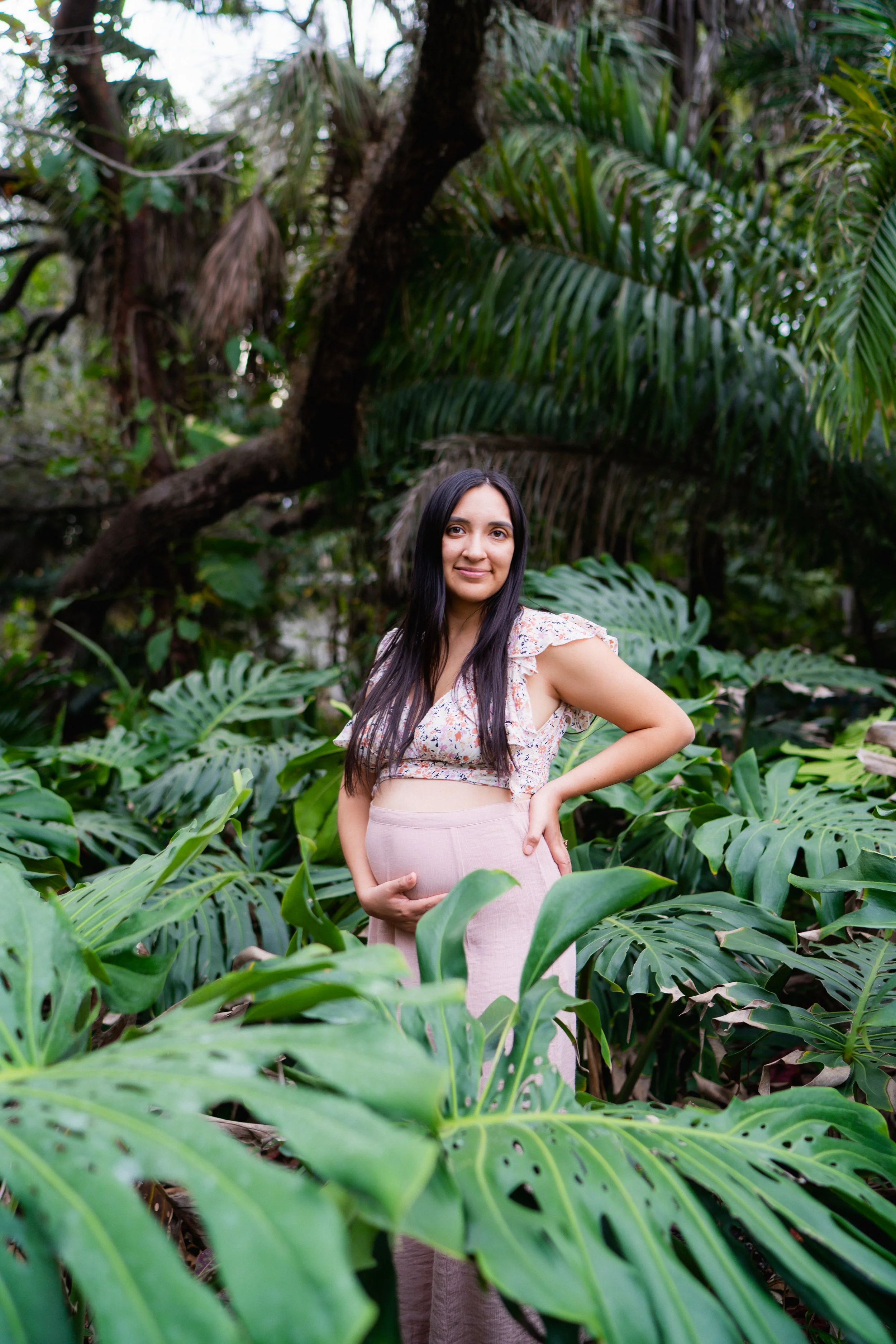 A woman standing in a lush green tropical garden, holding her pregnant belly with her right hand. She has long dark hair and is wearing a light-colored, floral-patterned top and beige pants.