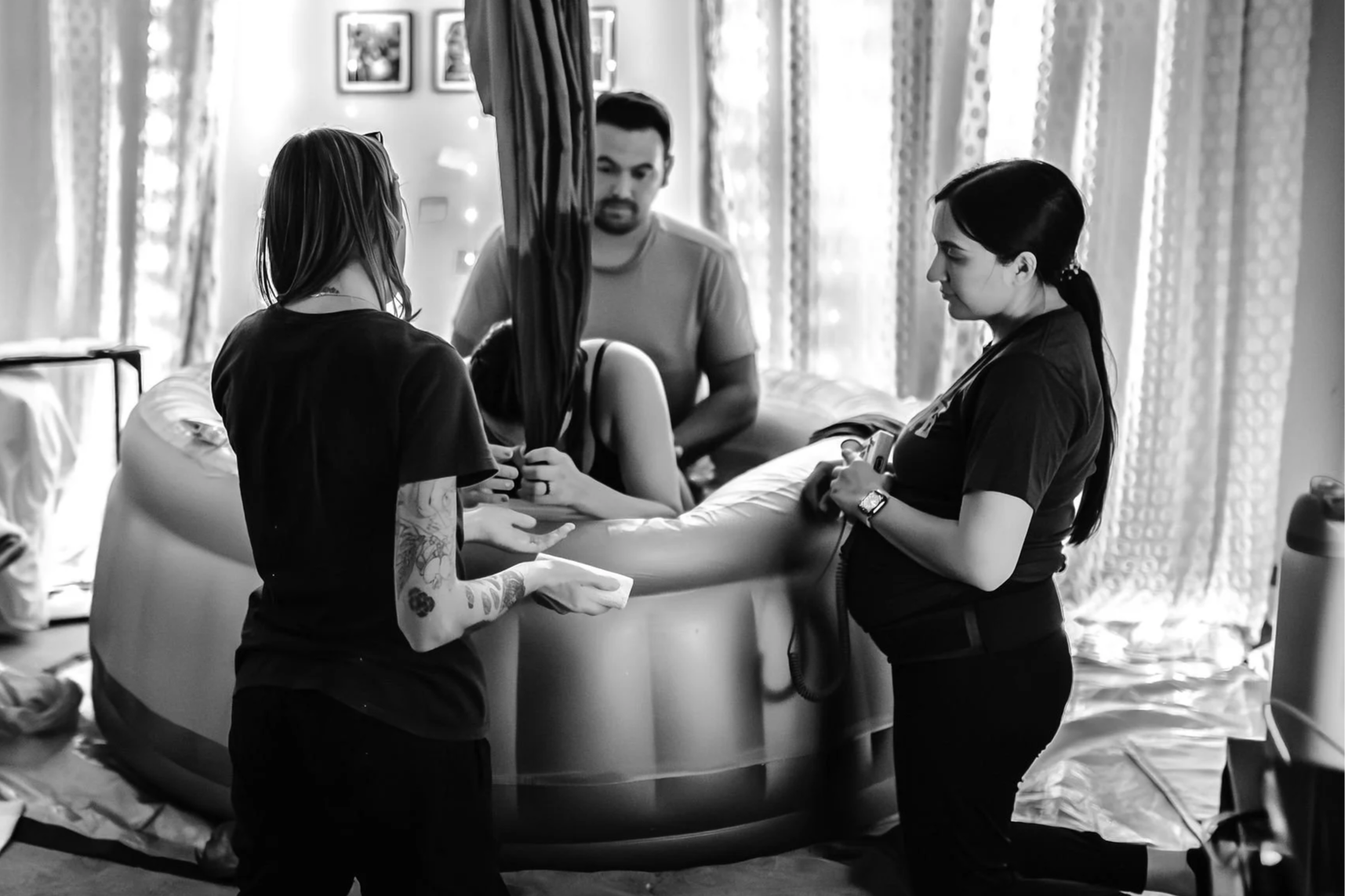 Group of people in a room with hospital bed, curtains, and artwork, with one person on the bed, two standing near the bed, and one in the background.