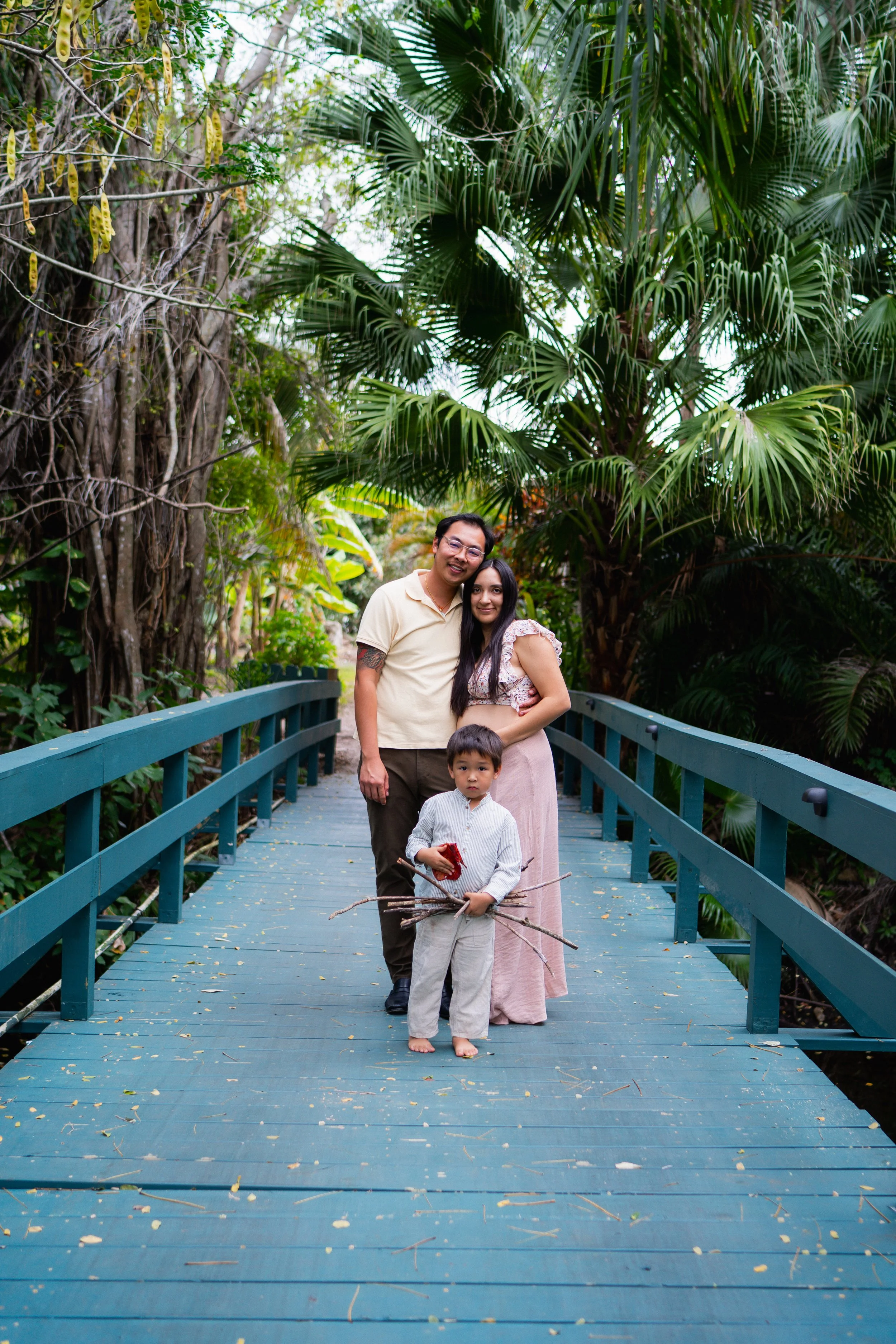 A family of four standing on a teal-colored bridge surrounded by lush green trees and tropical plants. The father, mother, and two children are smiling at the camera, with the mother and father standing close together and the children in front.