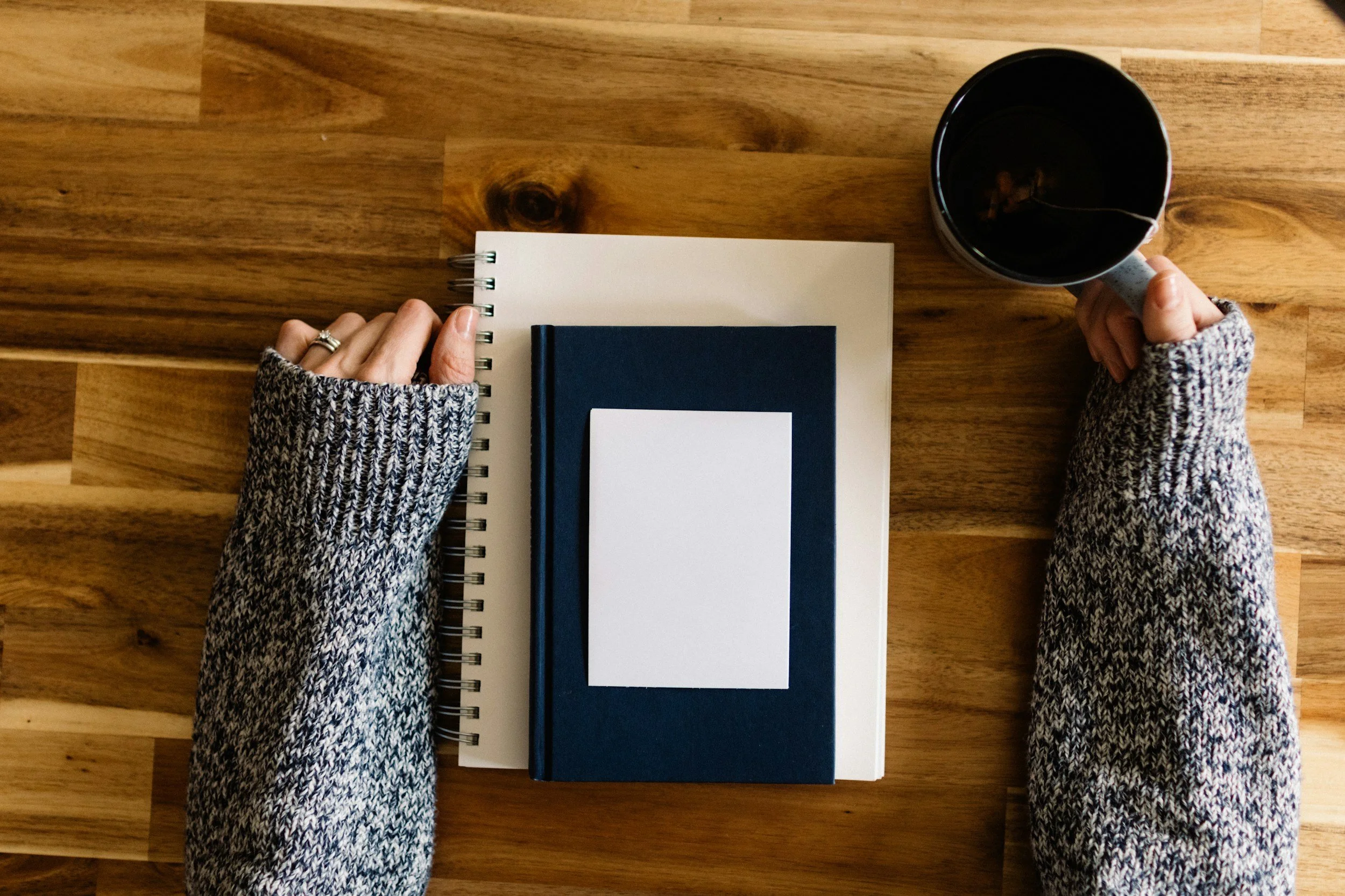 Overhead view of a wooden table with a person's hands, wearing a knitted sweater, holding a white coffee mug on the right side and a spiral notebook with a pen on the left side. The notebook's pages are blank, and the table has a natural wood grain finish.