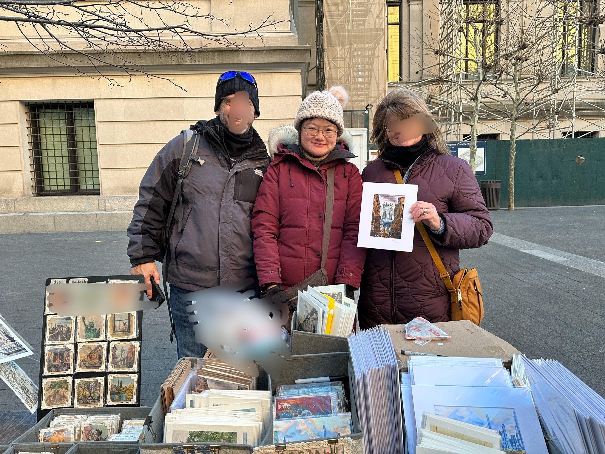 Three people standing behind a table of postcards and artwork at an outdoor street market. The person on the left is wearing a gray jacket, black hat, and sunglasses on hat. The person in the middle is wearing glasses, a beige knit hat with a pom-pom, and a maroon jacket. The person on the right is wearing a purple jacket with a black neck gaiter and holding a framed postcard of a city scene. Trees and a building with scaffolding are visible in the background.