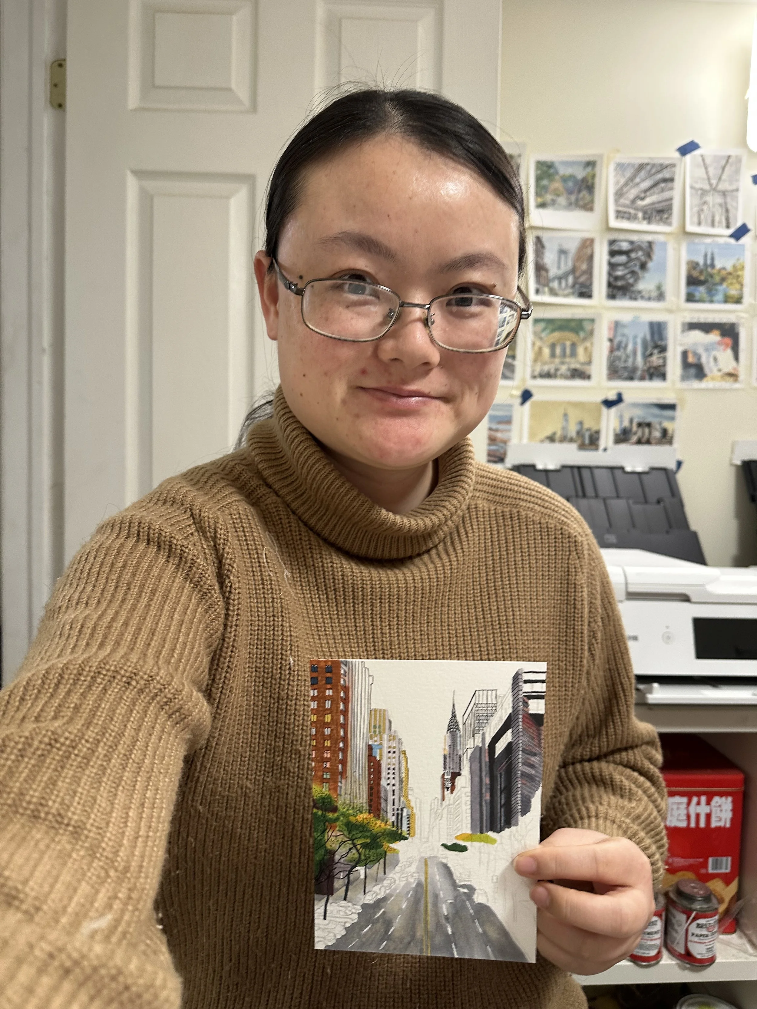 A woman wearing glasses and a brown sweater holding a painting of a city street with tall buildings and trees.