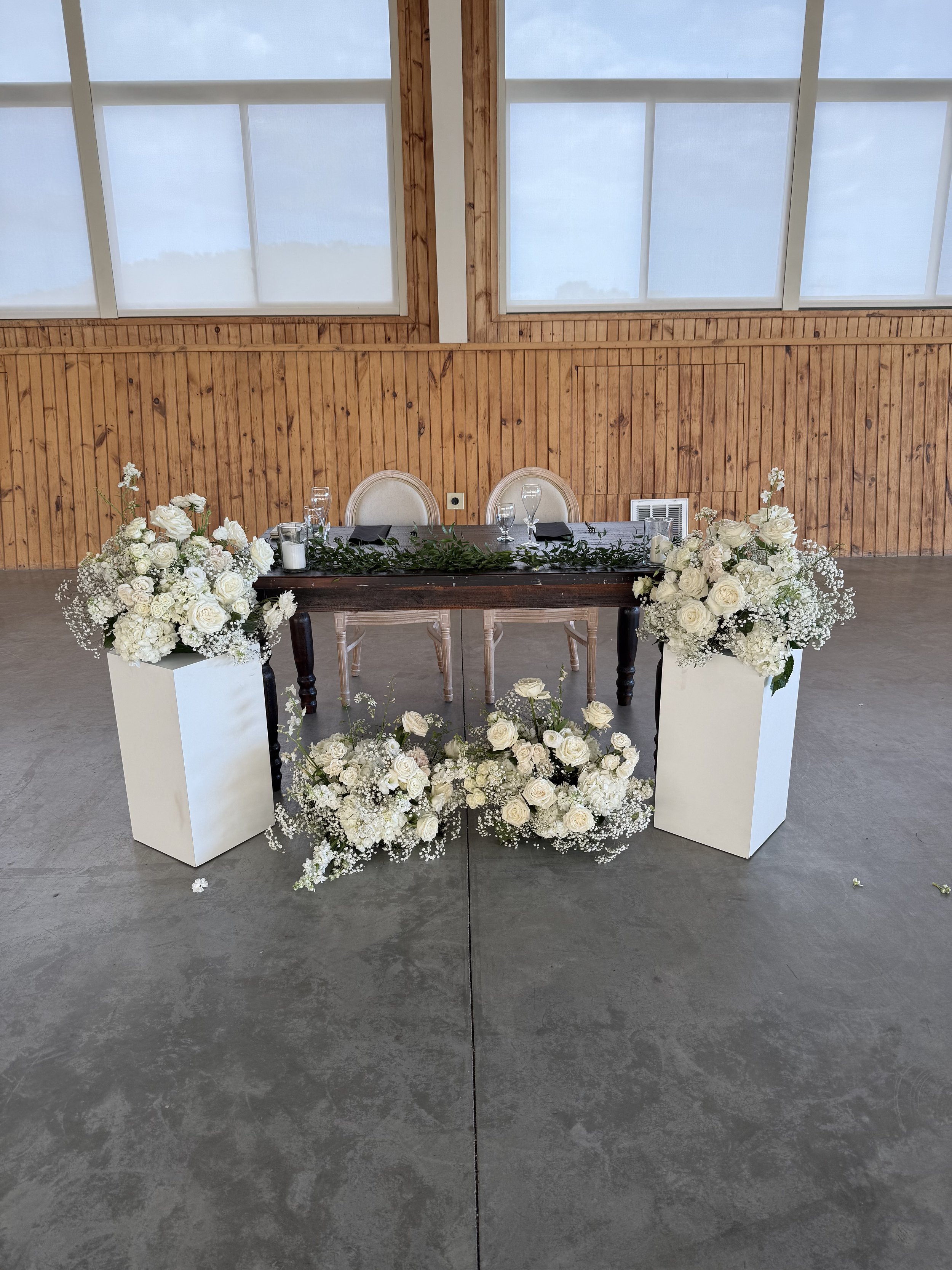 Table decorated with white flowers, candles, and glassware in front of a wooden wall with large windows.