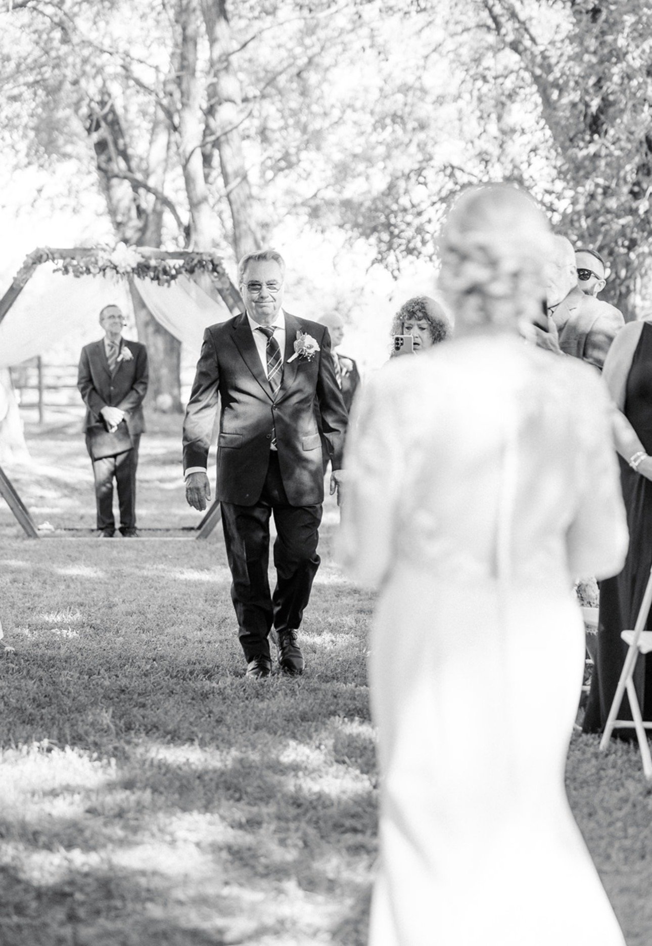 A black and white photo of a wedding ceremony outdoors with guests walking and a wedding arch in the background, trees, and grass visible.