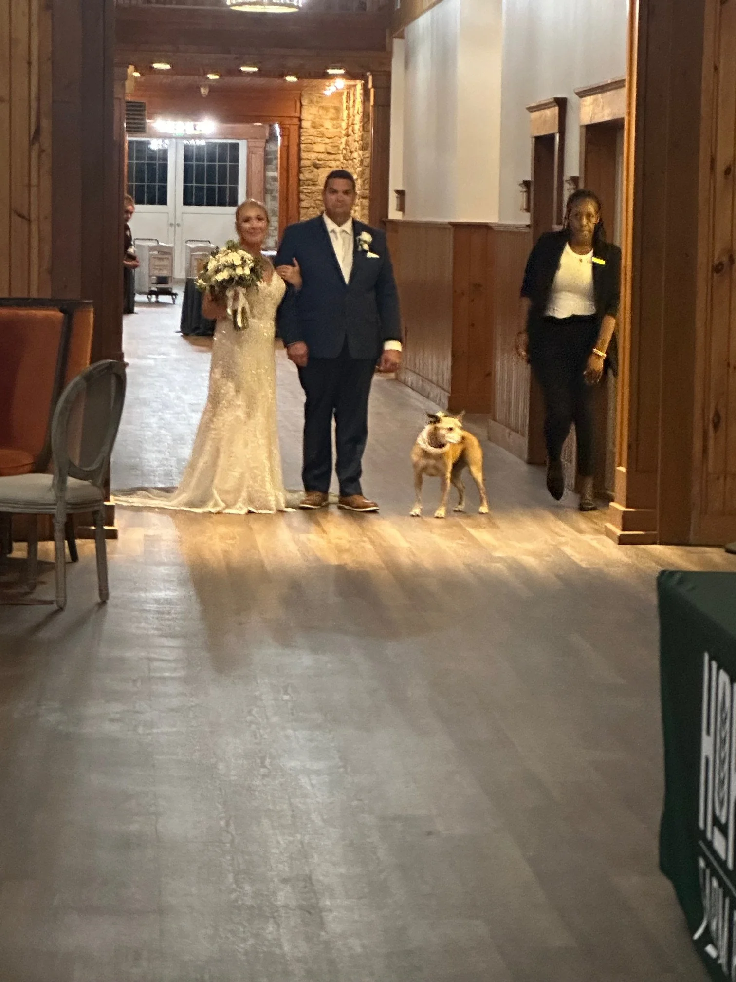 A bride in a white lace wedding dress holding a bouquet walking down a hallway with a man in a blue suit. An older woman, possibly a wedding planner or bridesmaid, walking beside them. A dog on a leash is walking ahead of the couple.