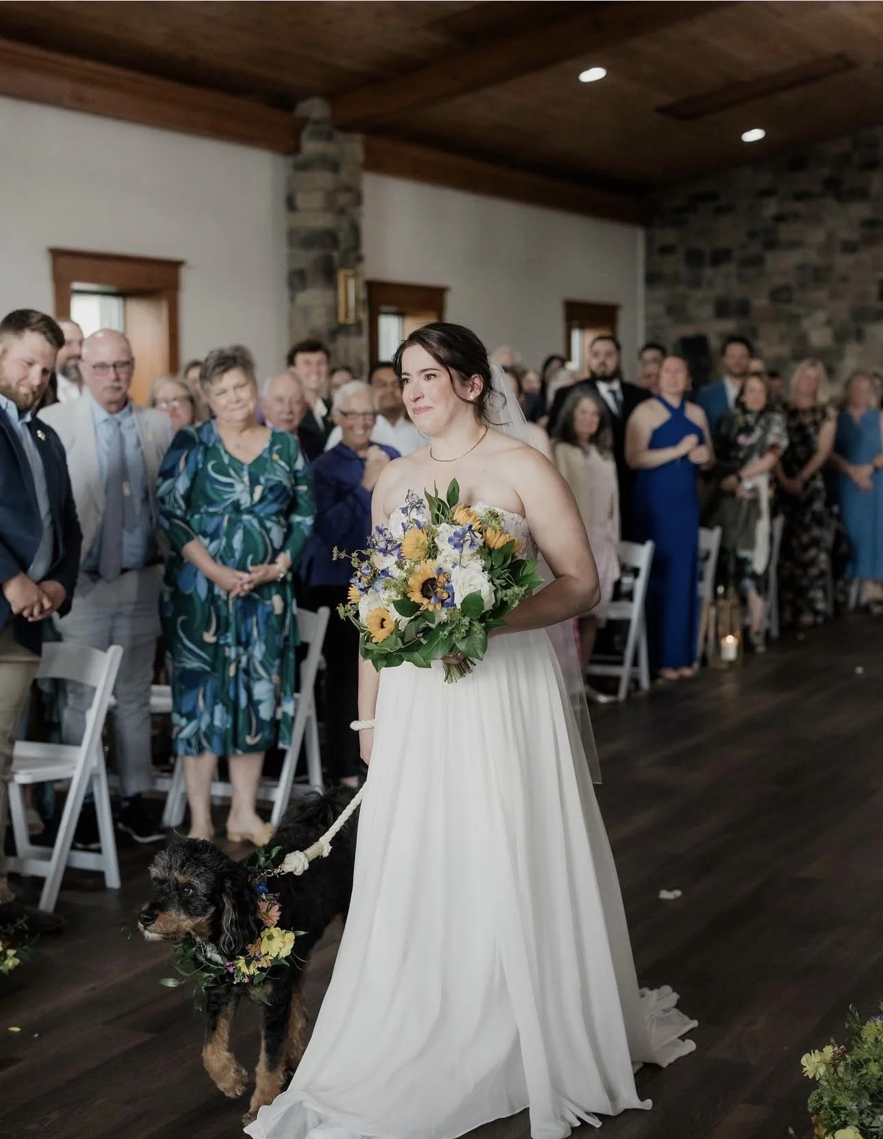 Bride in a strapless white wedding gown holding a bouquet of sunflowers, purple, and white flowers, walking a black dog decorated with a flower collar and leash, during a wedding ceremony with guests clapping and smiling in a rustic indoor setting with wooden ceiling and stone walls.