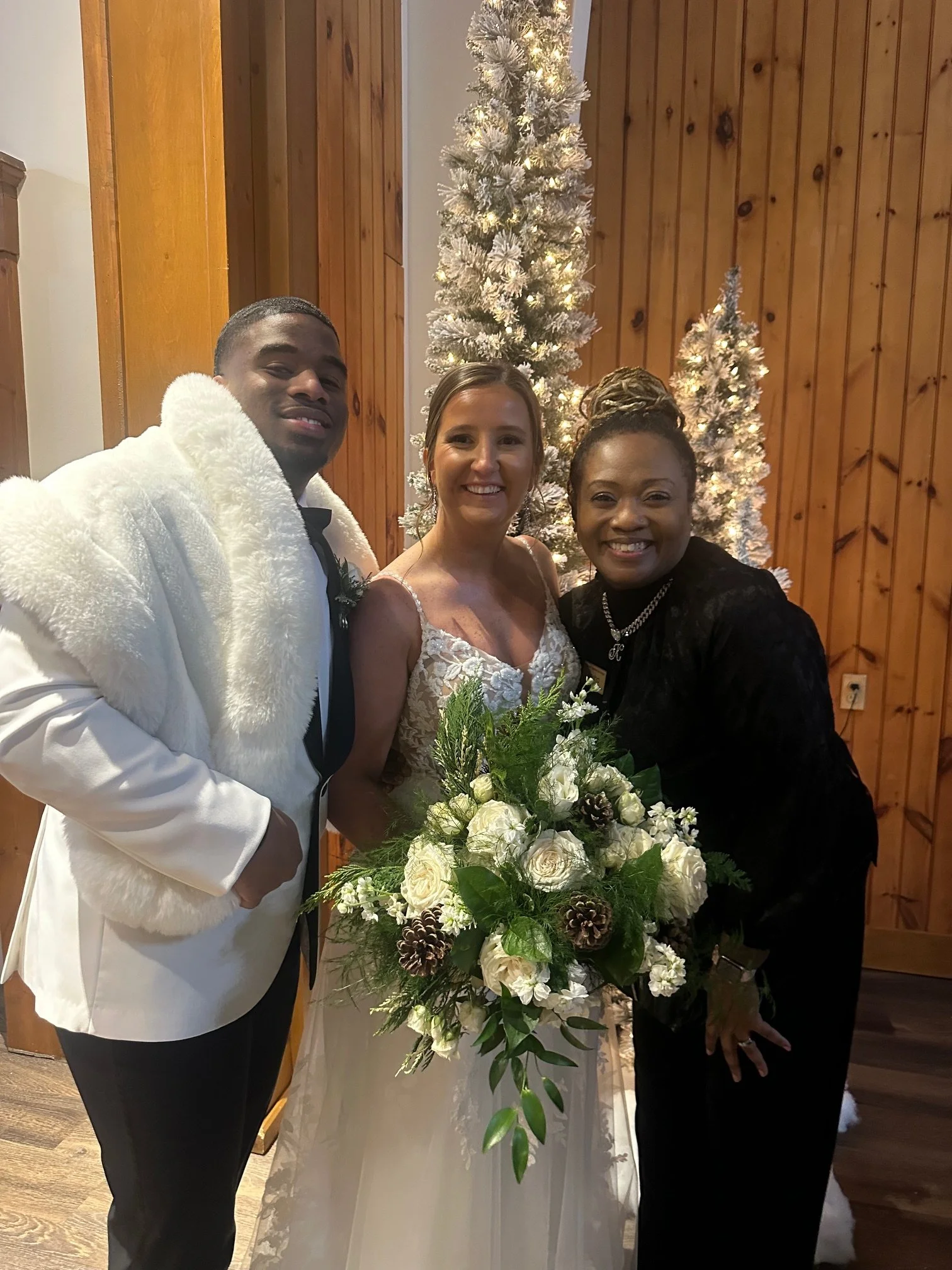 Three people standing together indoors, with a decorated Christmas tree in the background. The man on the left is wearing a white tuxedo jacket with a black bow tie, the woman in the middle is wearing a white wedding dress, and the woman on the right is wearing a black outfit with jewelry. They are smiling and holding a large floral bouquet.