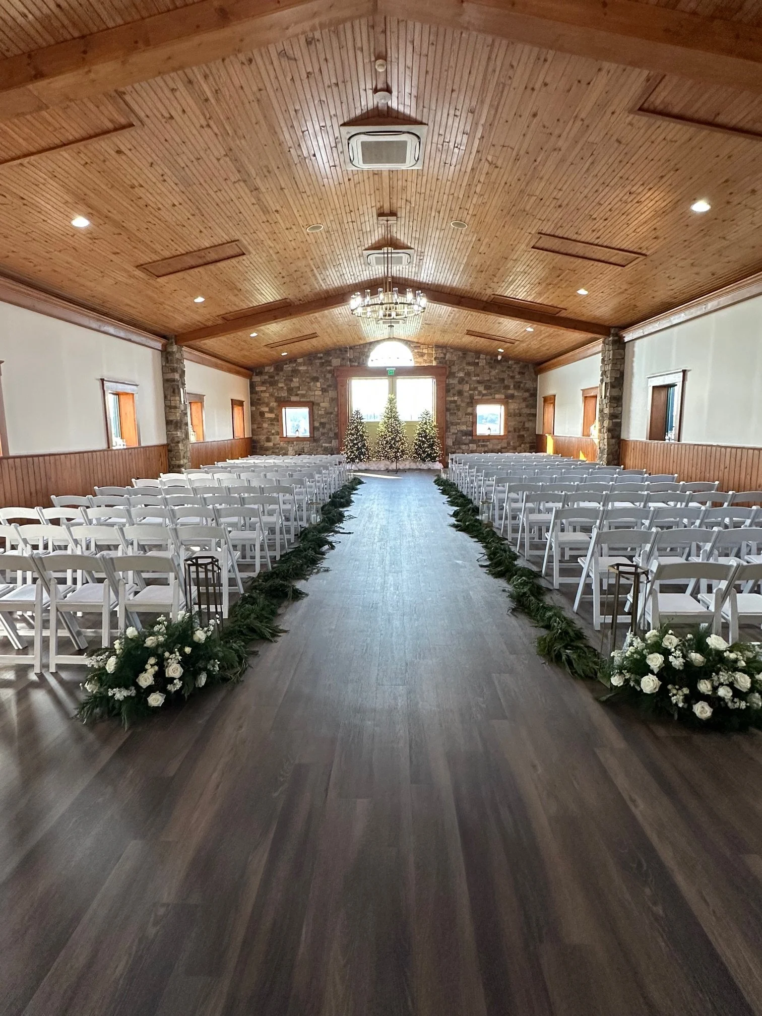 Interior of a wedding venue with rows of white chairs, a central aisle decorated with greenery and white flowers, and a backdrop with three decorated Christmas trees and a large window.