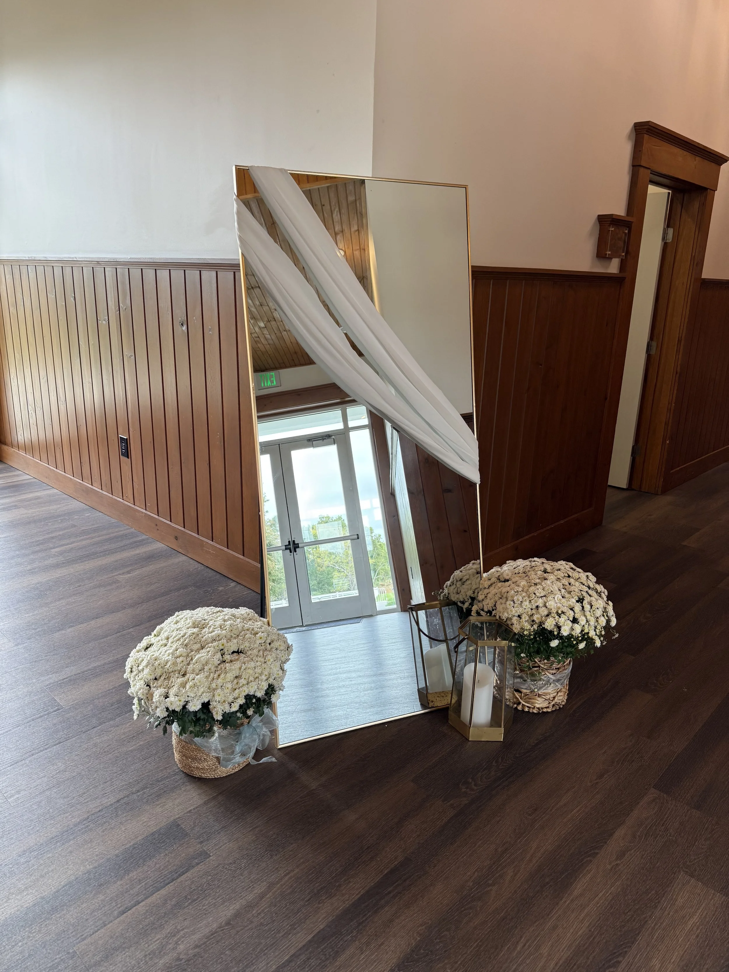 A large, standing mirror reflects a doorway and greenery outside. Two baskets of white chrysanthemums are on either side of the mirror, and a lantern with candles is on the right.