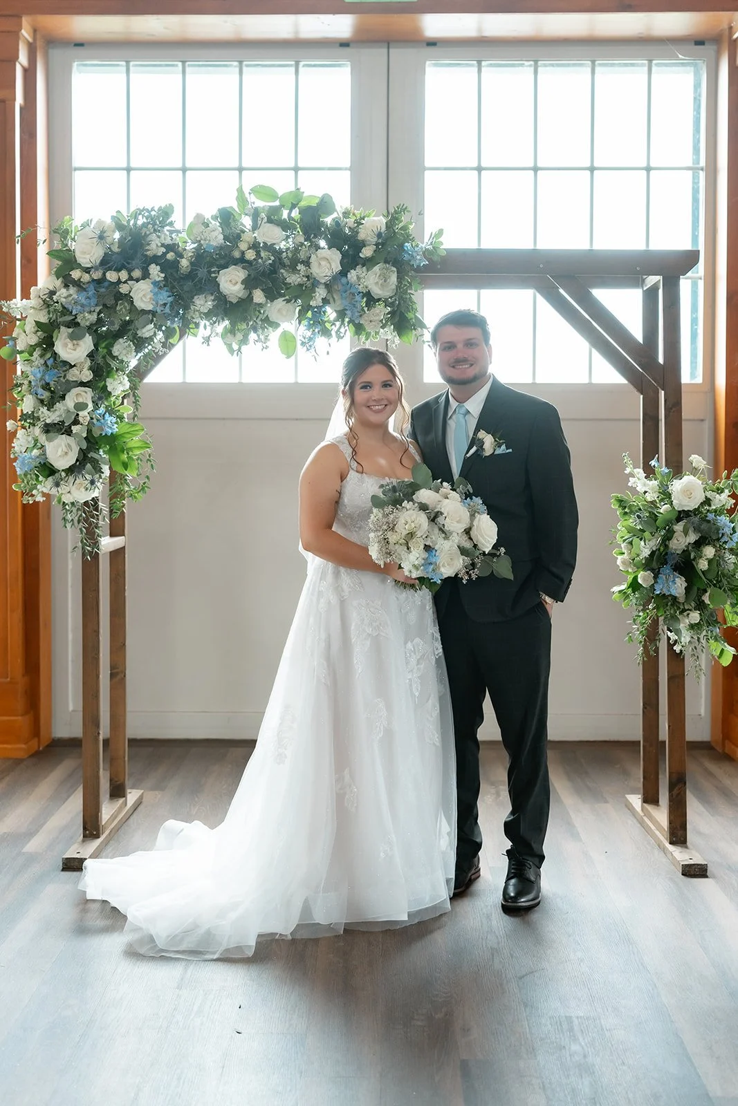 A bride and groom standing under a flower arch at their wedding, smiling at the camera.