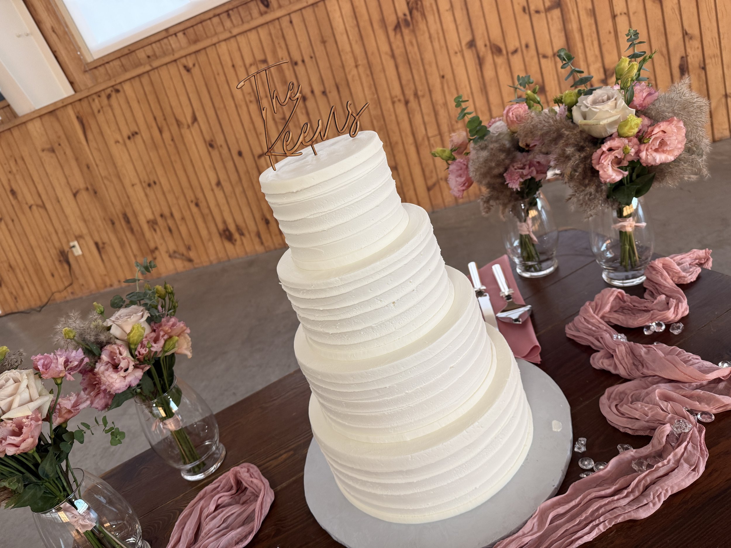 A wedding cake with white icing and a topper that says 'The Jess's,' surrounded by pink and white flowers in glass vases, on a wooden table draped with pink fabric, against a wood-paneled wall.
