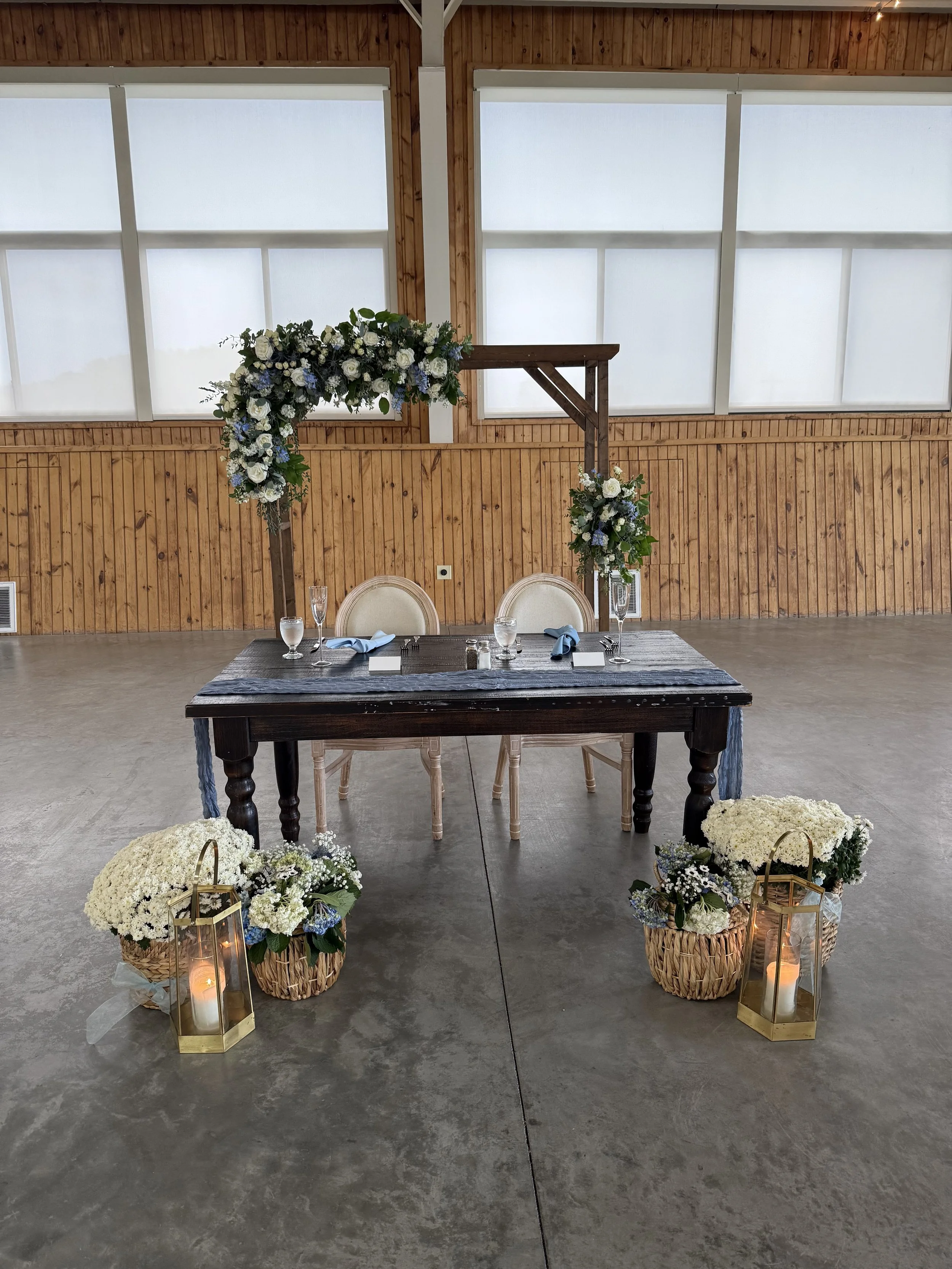 A wedding setup with a wooden table and floral arrangements, including lanterns and candles, on a concrete floor against a wooden paneled wall with large windows.