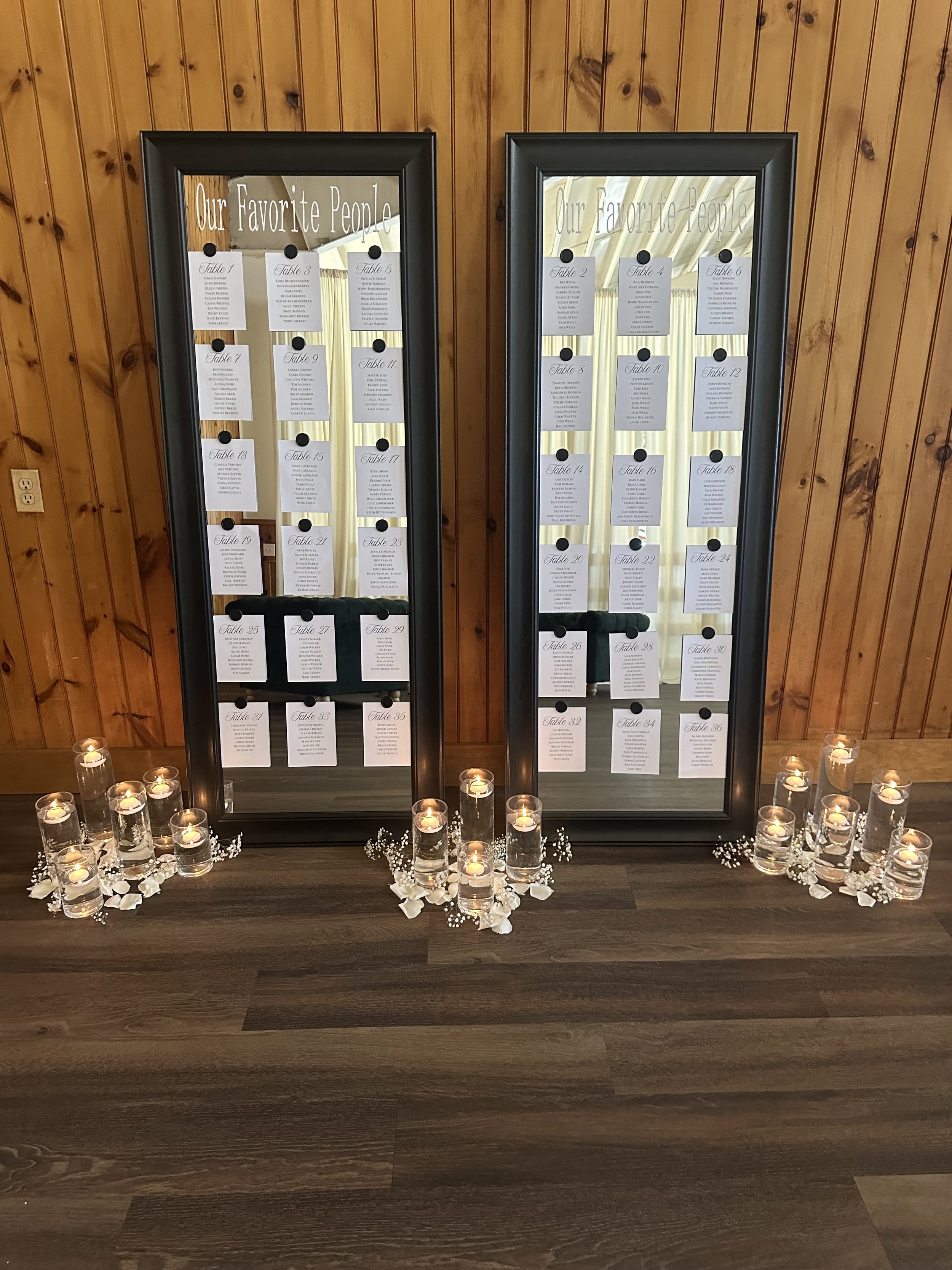 A mirrored display with 'Our Favorite People' written at the top, showing table assignments for a wedding or event, decorated with candles, rose petals, and small white flowers on a wooden surface, against a wooden-paneled wall.