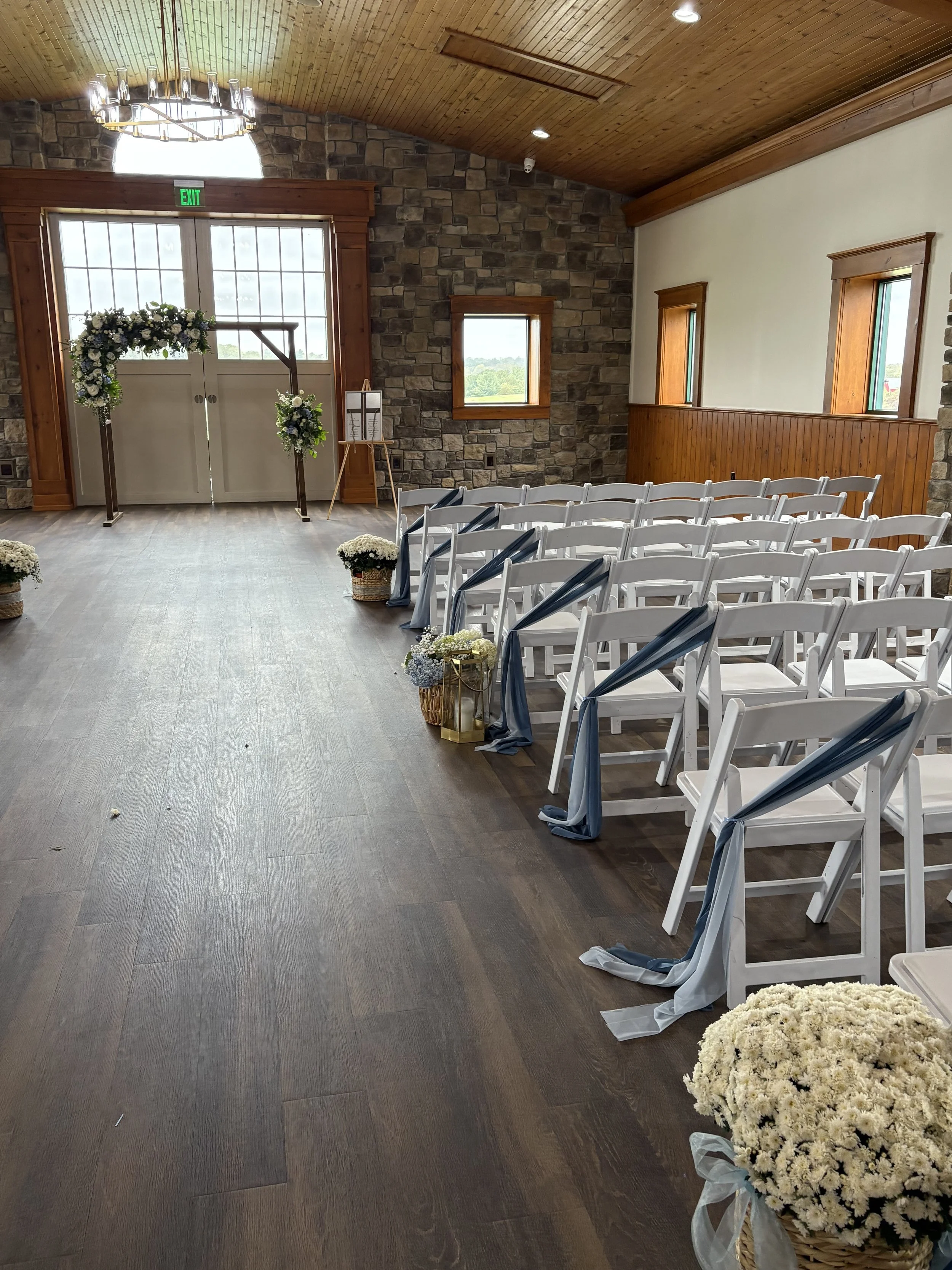 Wedding ceremony setup with white chairs, floral arrangements, a decorative arch with flowers, and a wooden door at the front, inside a rustic venue with stone and wooden walls.