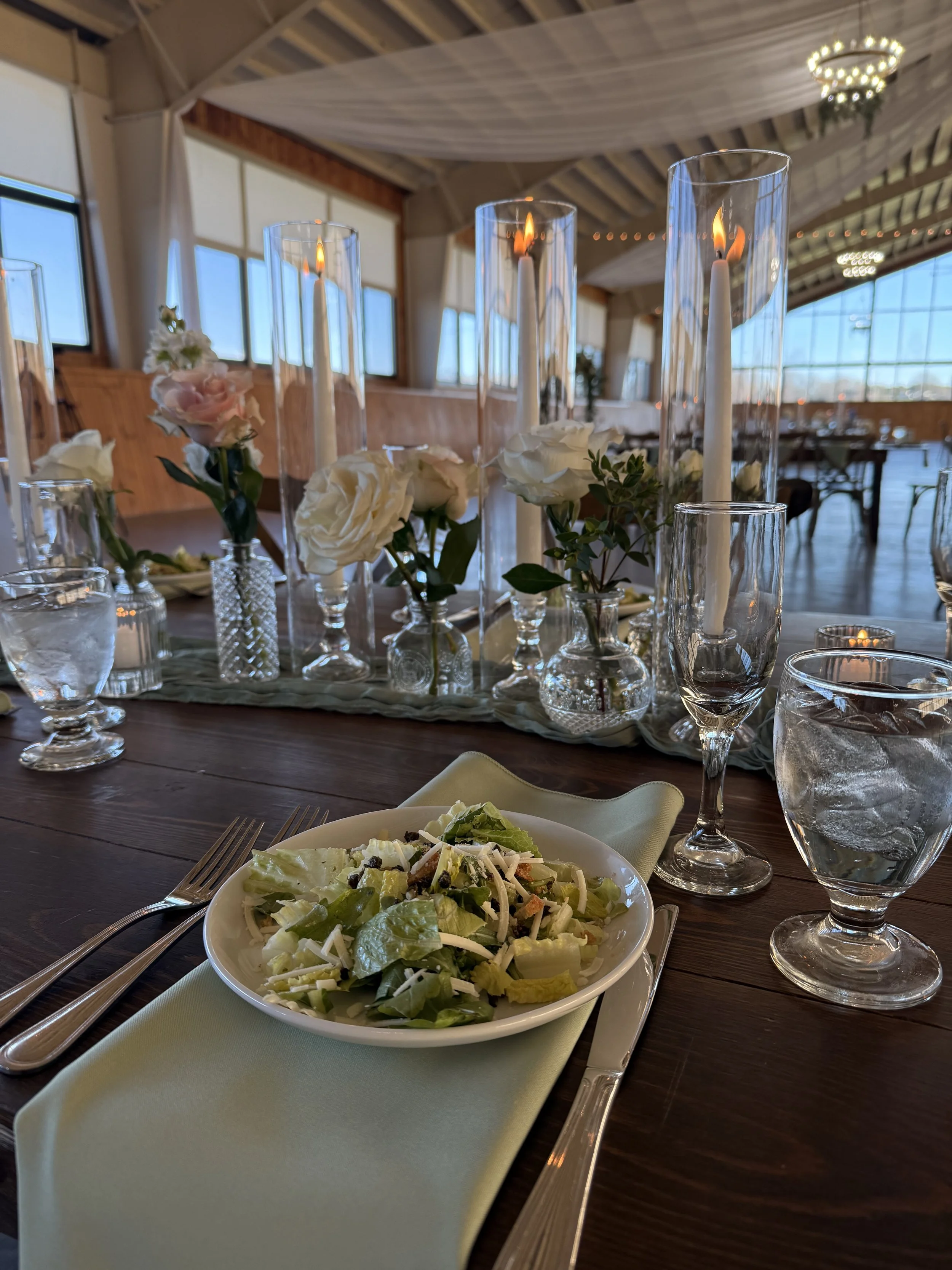 Elegant dining table with a bowl of green salad, surrounded by water glasses, wine glasses, and candleholders with lit candles, set in a decorated event space with high ceilings, large windows, and chandeliers.