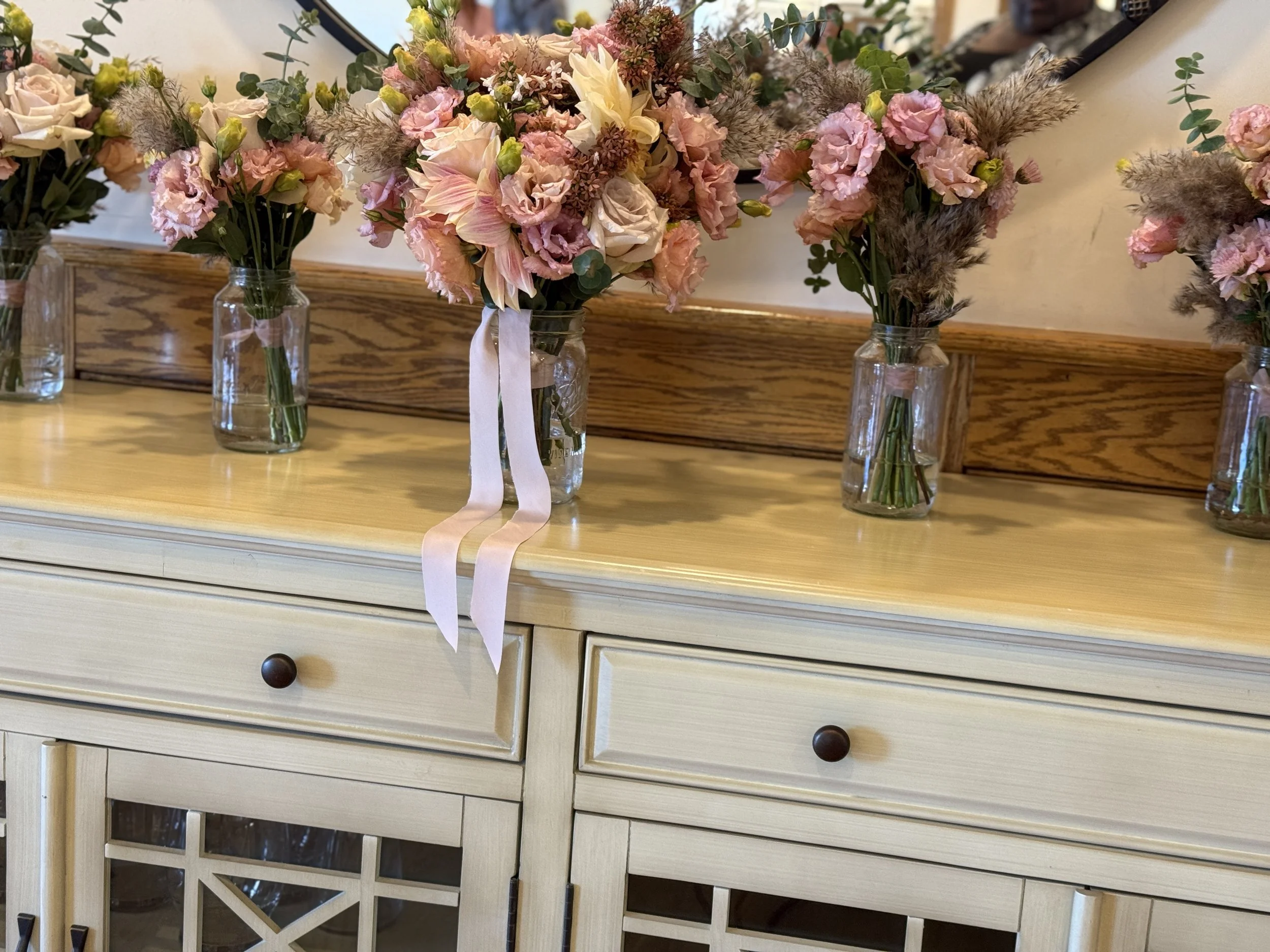 A wooden dresser with multiple drawers and dark round knobs, decorated with multiple glass vases filled with pastel-colored flowers, including pink, cream, and light yellow flowers with greenery, on top of the dresser.
