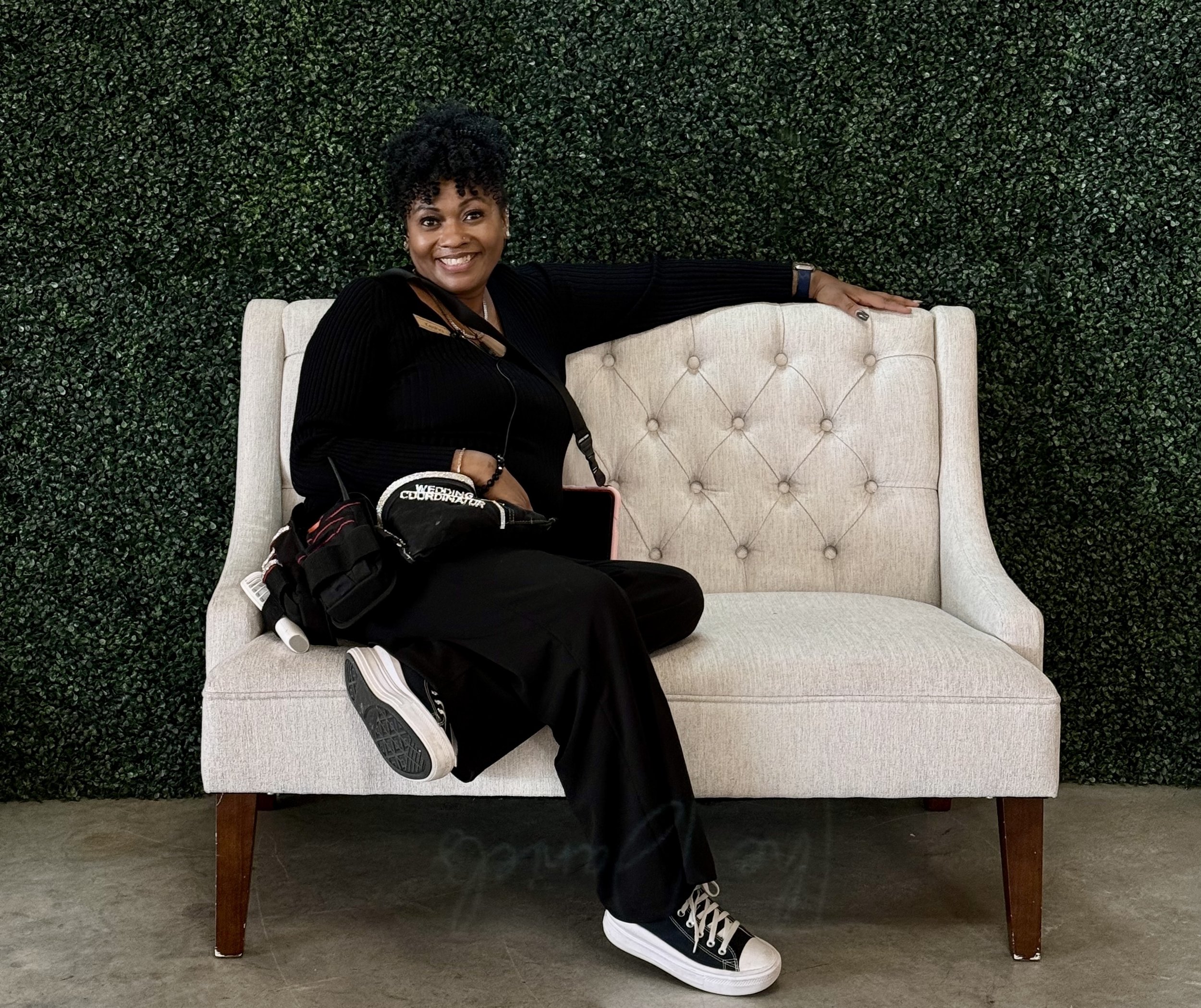 Woman sitting on a beige tufted armchair against a green hedge background, smiling, wearing a black top, black pants, black and white sneakers, and holding a black bag.