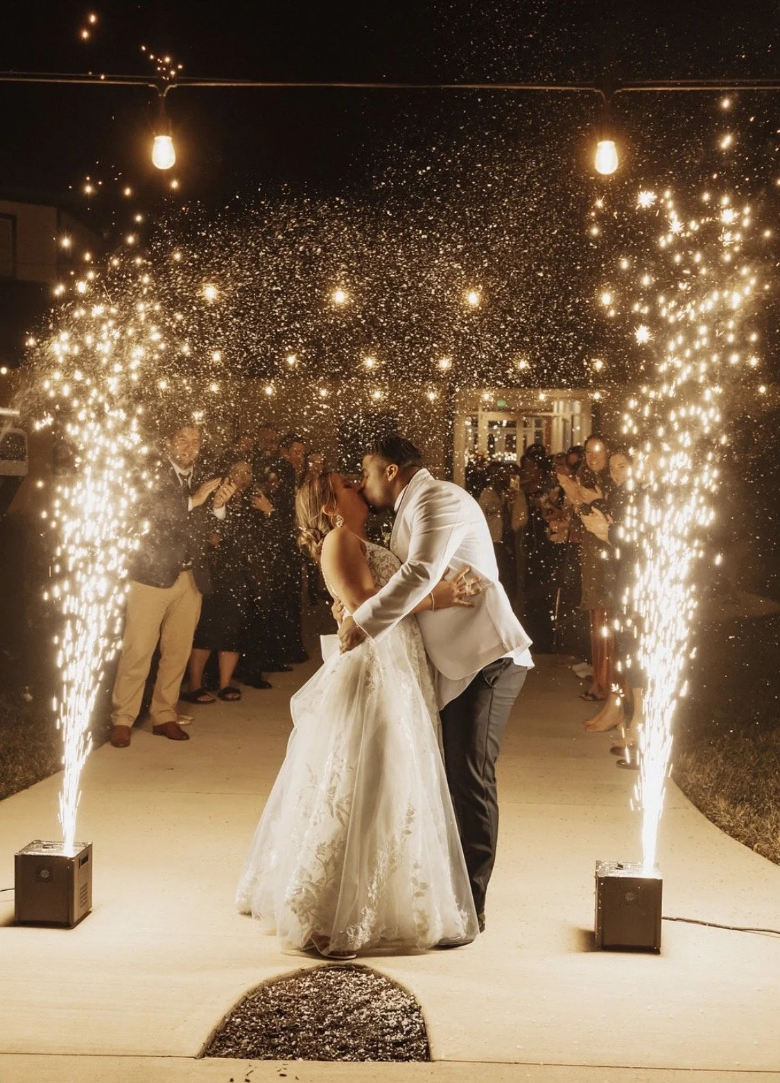 Bride and groom kissing under fireworks during their wedding celebration at night, with guests watching in the background.