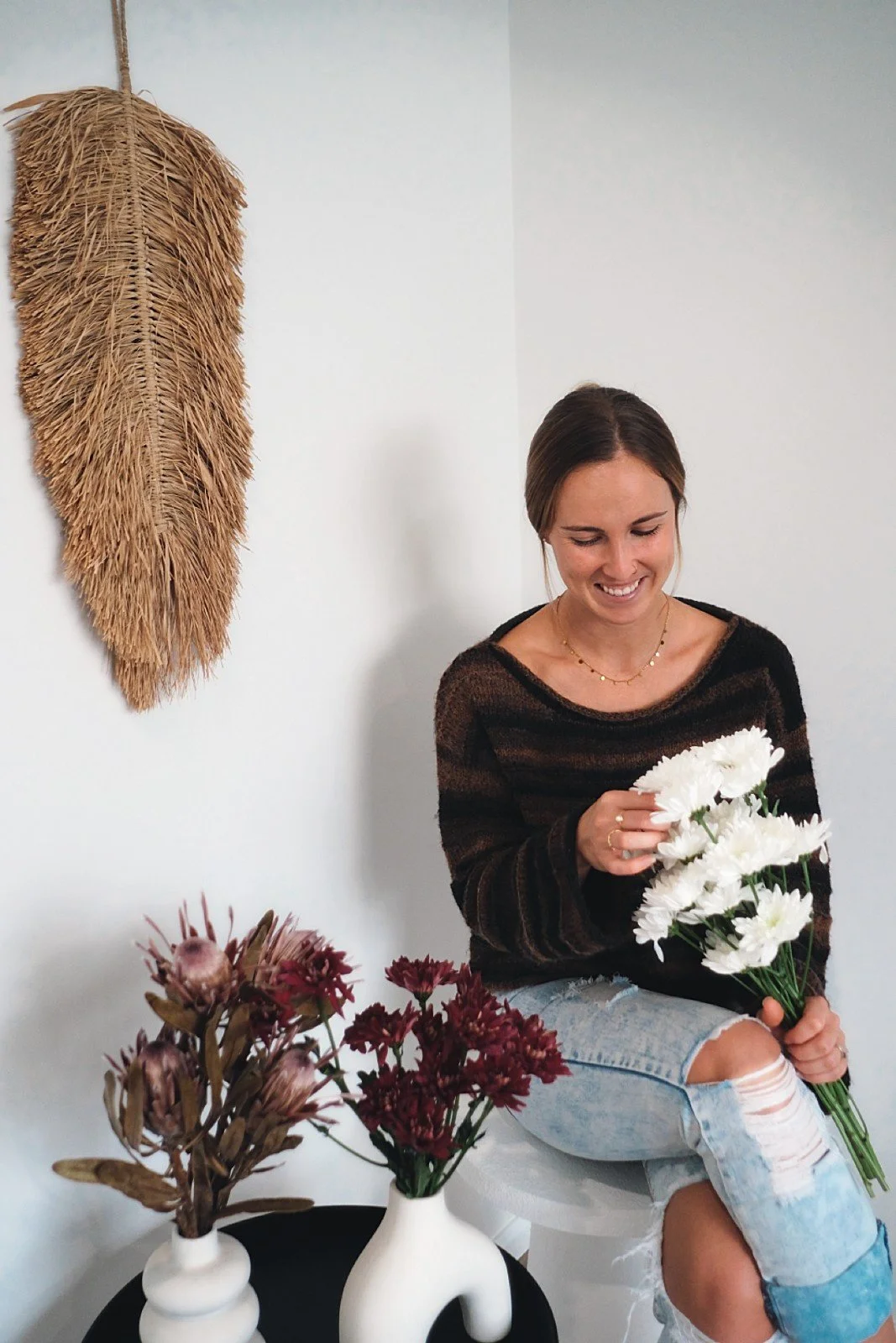 Kirstie Shields sitting on a white stool, smiling and holding a bouquet of white daisies, with a wall and a woven wall hanging in the background, and vases with dried flowers on a black table, from Salt & Bloom Co
