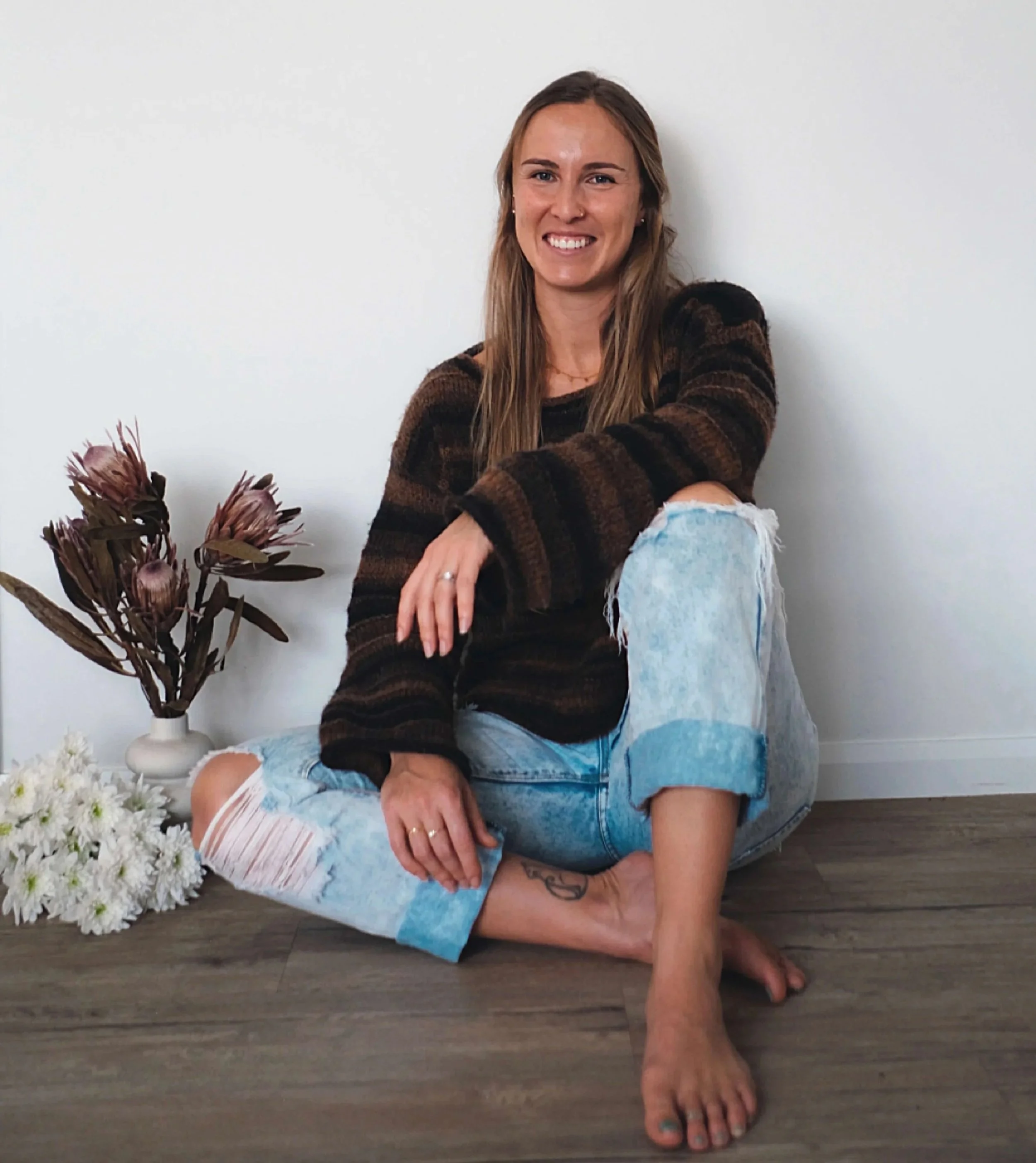 Kirstie Shields with long brown hair, smiling and showing her teeth, sitting on the floor with her knees up, wearing a striped brown and black sweater and ripped blue jeans, from Salt & Bloom Co