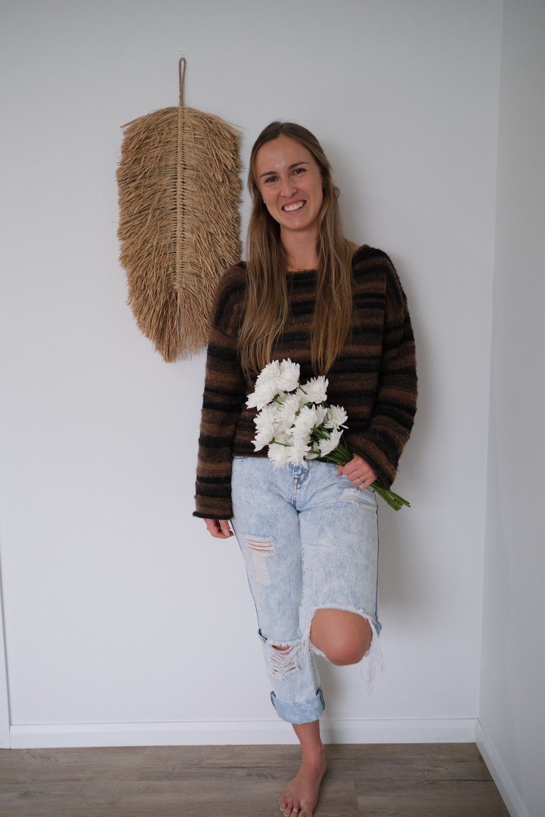 Kirstie Shields with long brown hair holding white flowers, wearing a striped dark brown and black sweater and light-wash ripped jeans, standing barefoot against a white wall with a large straw decoration hanging behind her from Salt & Bloom Co