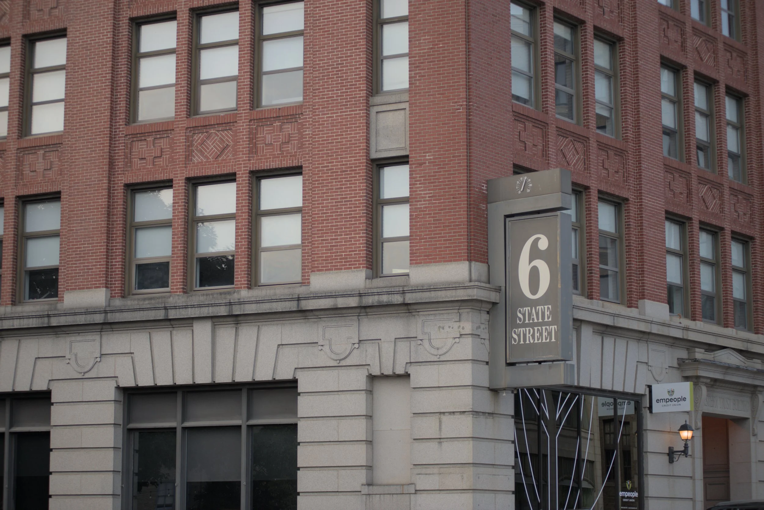 A red brick building with multiple windows and decorative brick patterns, a street sign displaying '6' and 'STATE STREET', and part of a lamppost at street level.