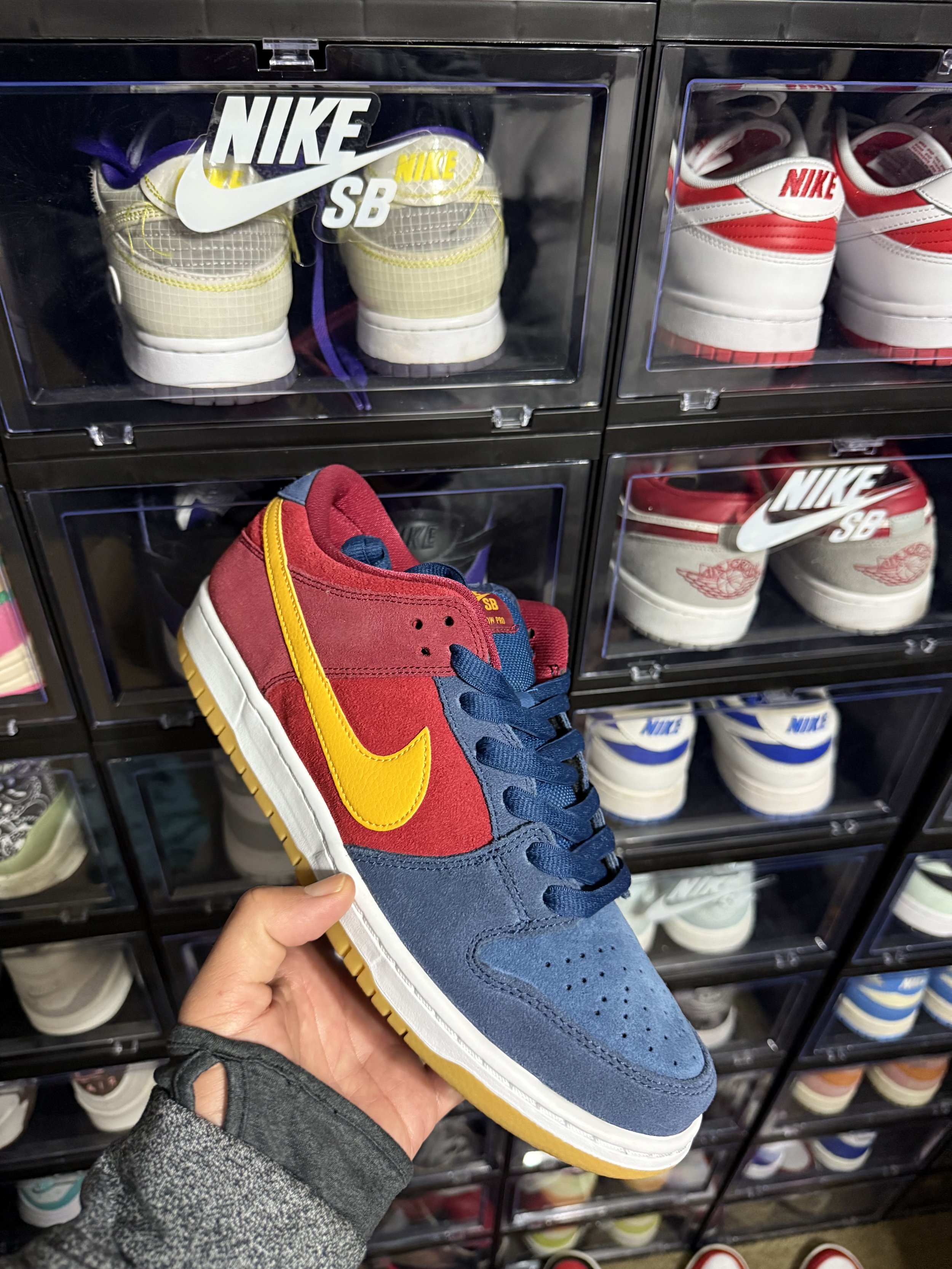 A person holding a multicolored Nike SB sneaker with a yellow Nike swoosh, navy blue accents, and a white rubber sole in front of a glass shoe display case filled with various Nike sneakers.