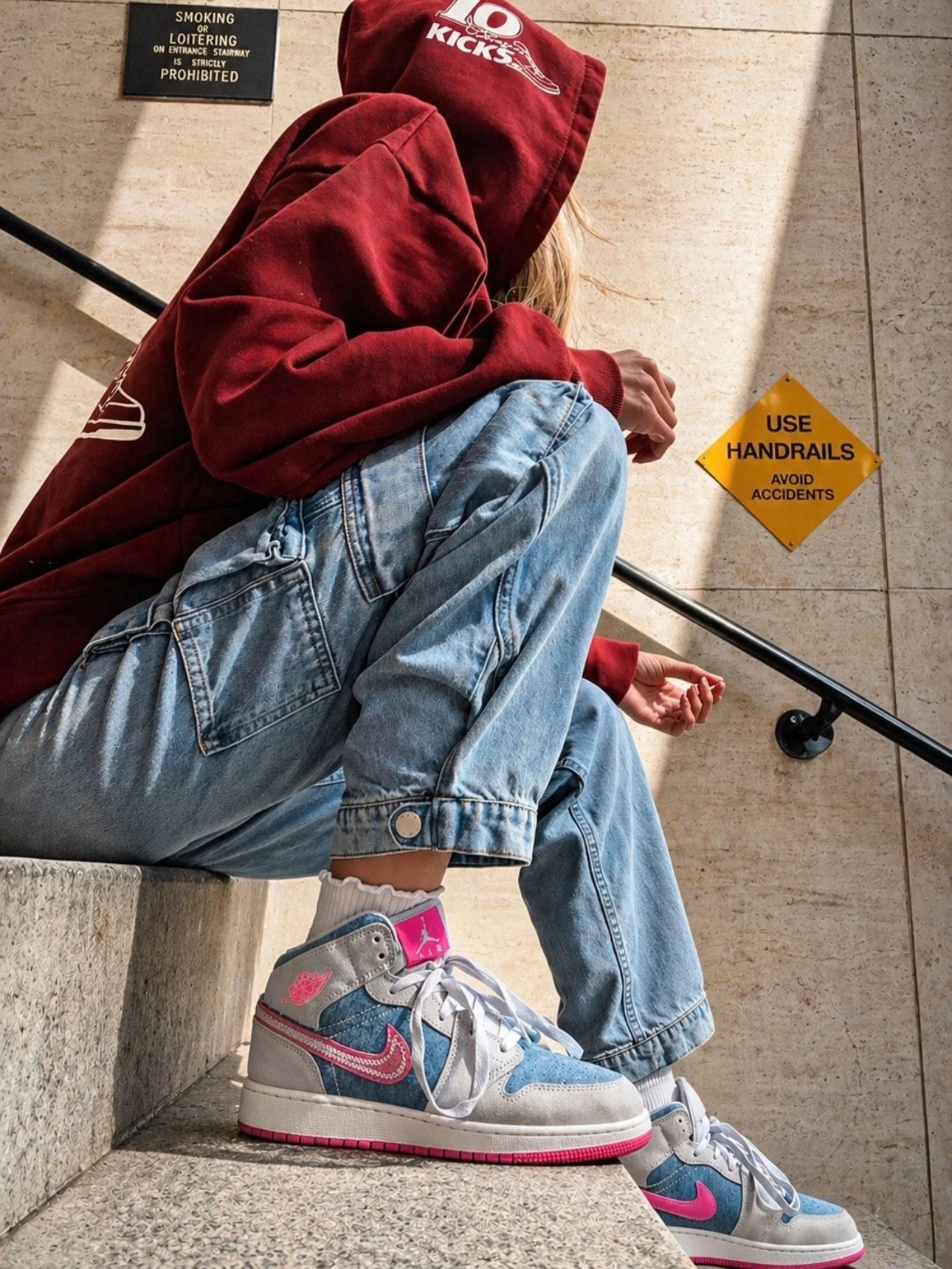 A person sitting on stone stairs, wearing a red hoodie, light blue jeans, and Air Jordan sneakers with pink and blue accents. Their face is hidden under the hoodie.