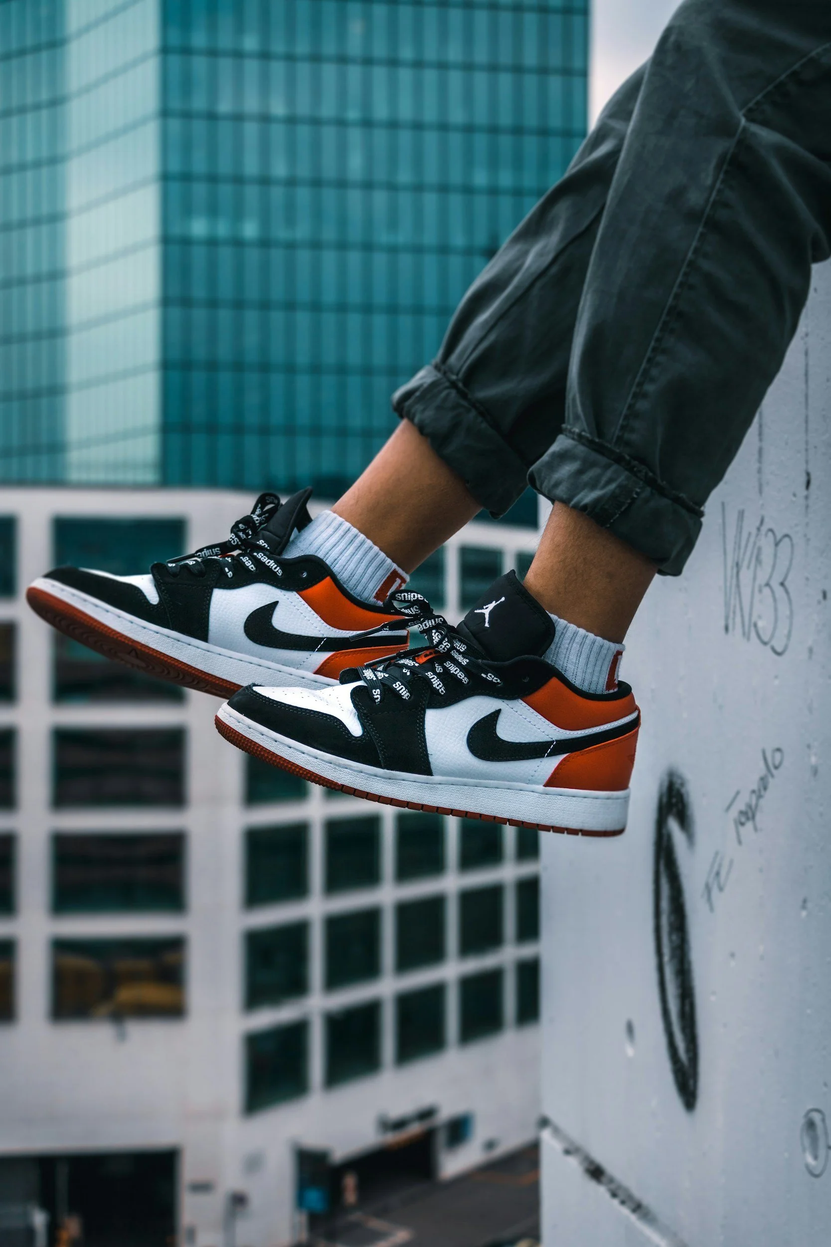 Close-up of a person's feet wearing Nike sneakers and gray socks, hanging off a graffiti-covered ledge in an urban area with modern glass buildings in the background.