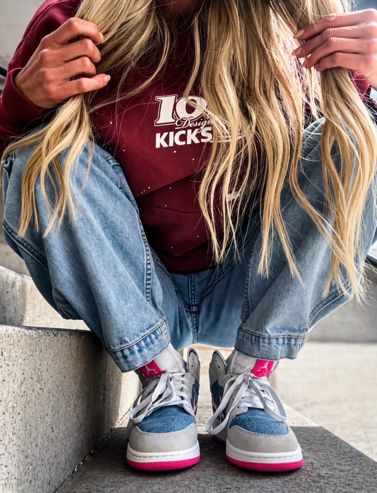 Person wearing a maroon sweatshirt with a logo, blue jeans, white sneakers with pink accents, sitting on concrete stairs, holding their hair, with long blonde hair.