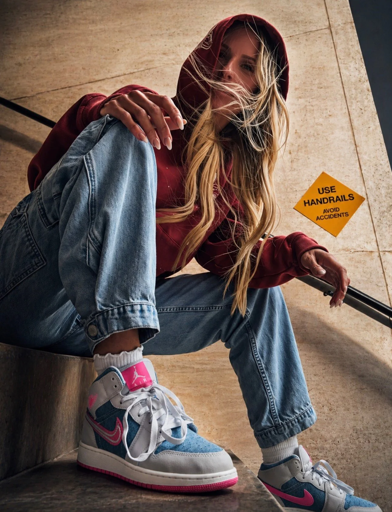 A woman with long blonde hair wearing a red hoodie, blue jeans, and colorful Air Jordan sneakers sitting on a bench in an indoor space, with a yellow sign in the background that says 'Use Handrails, Avoid Accidents.'