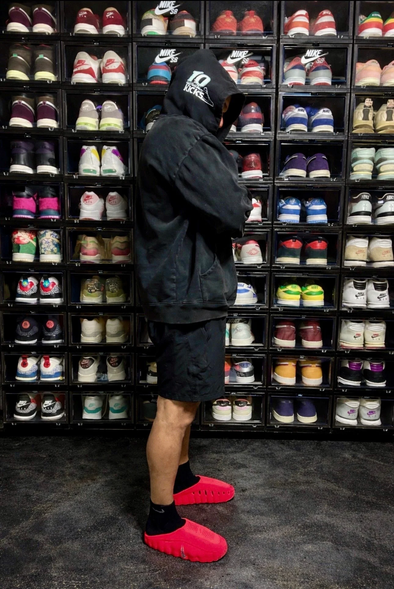 Person in black hoodie and shorts trying on a pair of large red sneakers at a shoe store with wall of colorful sneakers in display cases behind him.