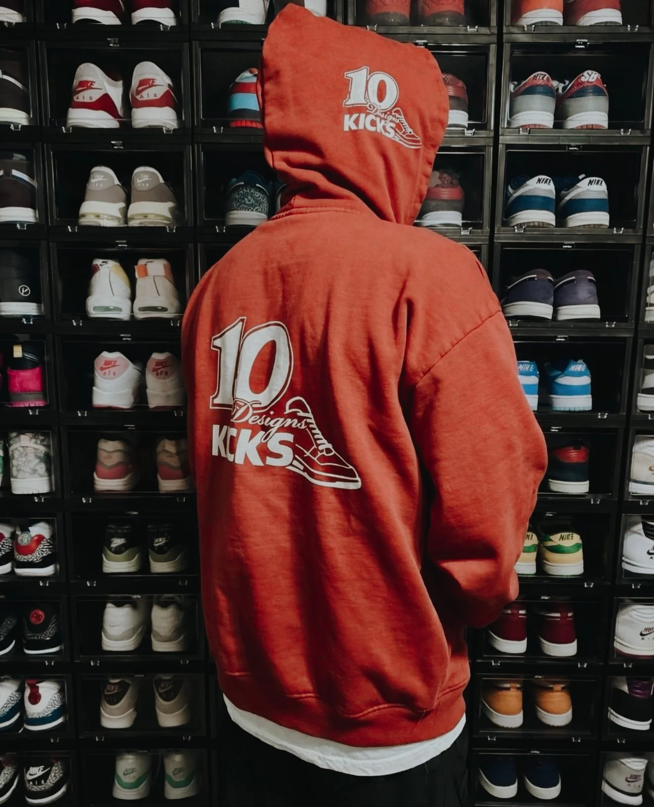 Person wearing a red hoodie with a logo that says '10 Kicks' and a sneaker graphic, facing shelves filled with various sneakers.