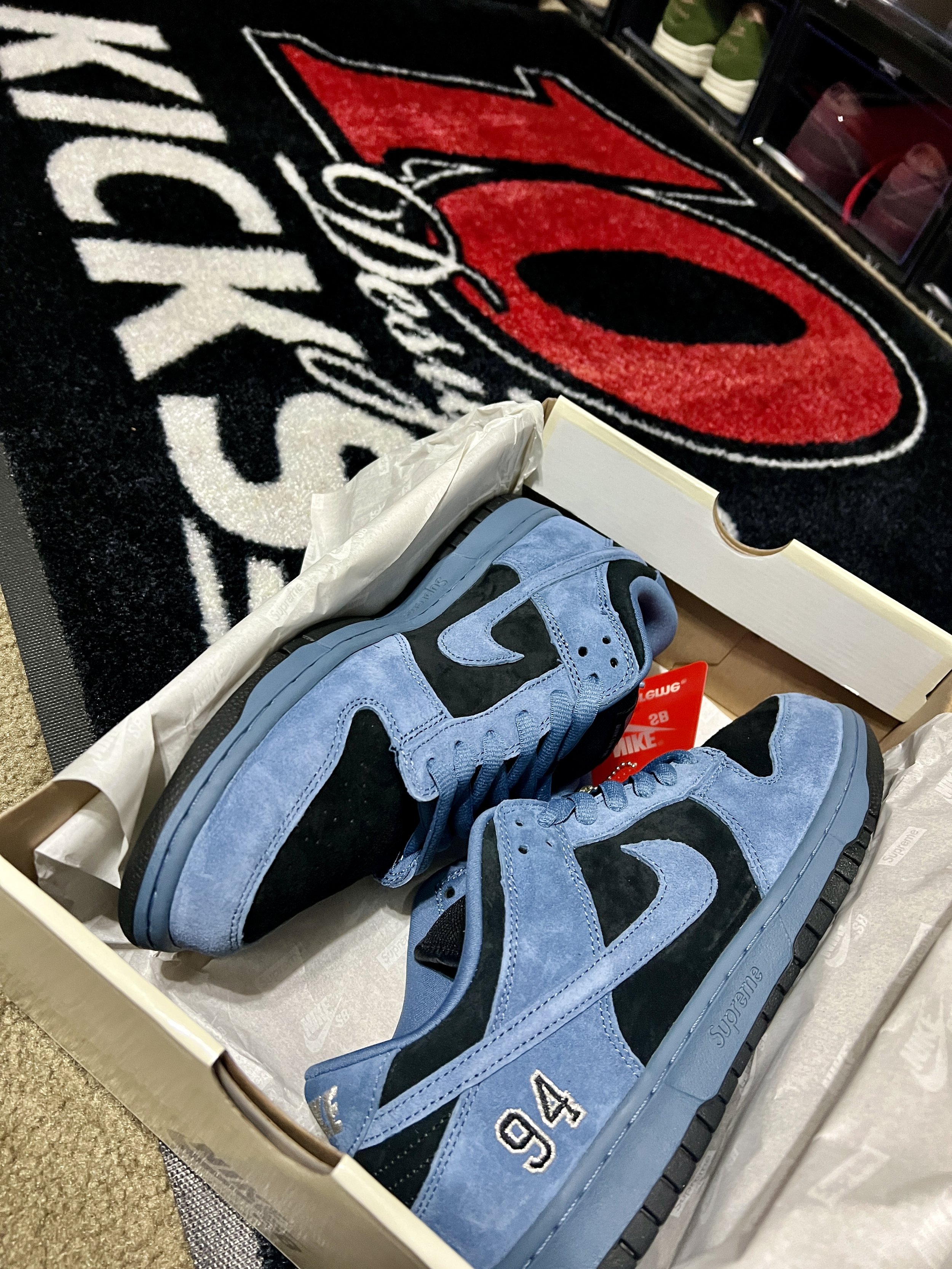 A pair of Nike sneakers in a box, with light blue suede and black accents, including the number '94' embroidered on the side, placed inside the shoe box with tissue paper. There is a black and red rug with the logo of the Arizona Cardinals football t