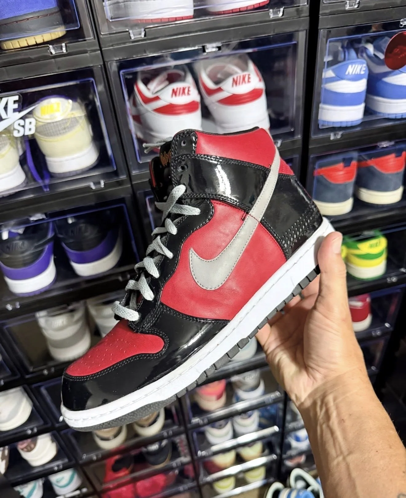 Person holding a red, black, and gray Nike sneaker in front of a display of various other sneakers stored in lockers.