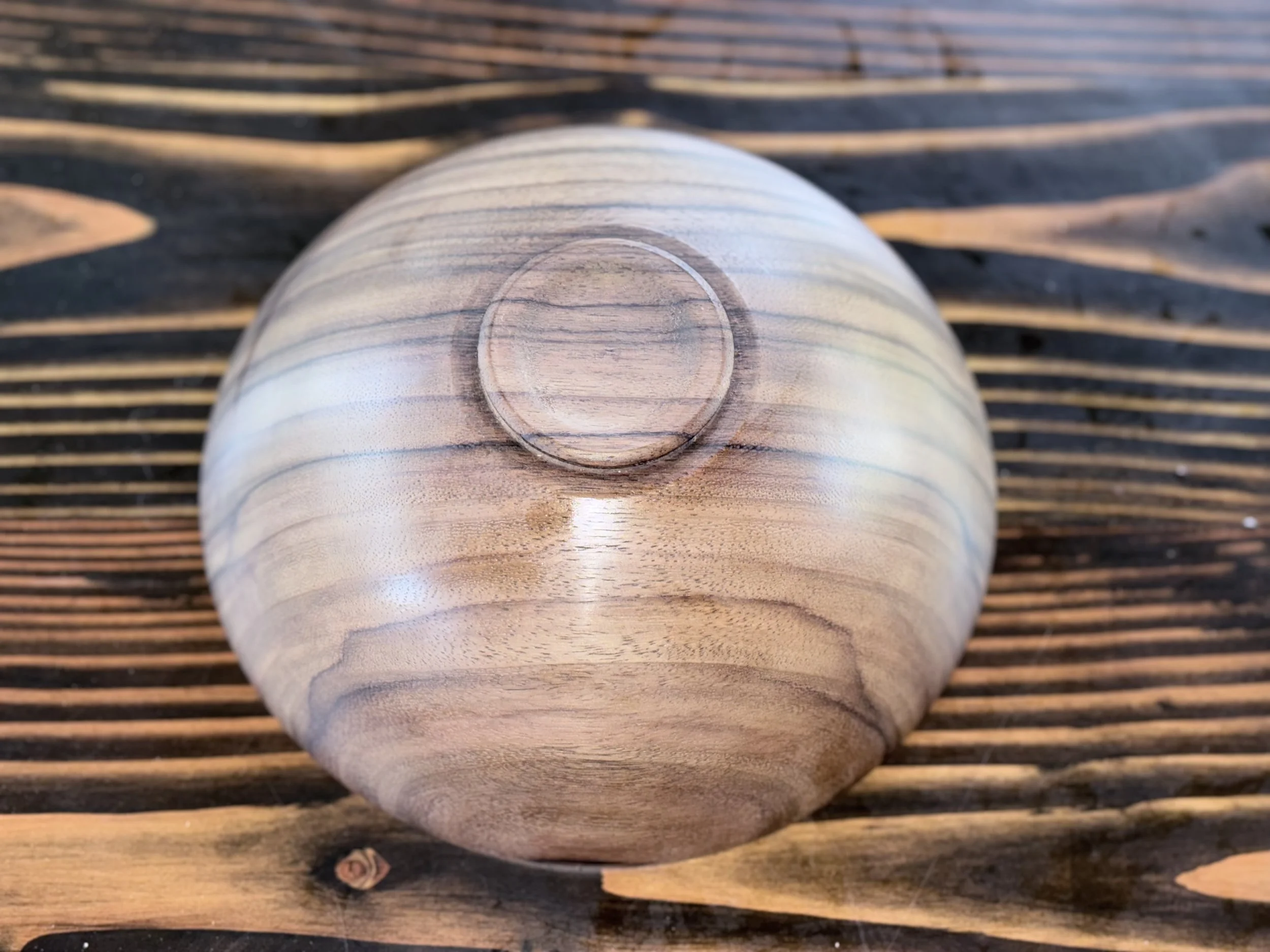 Close-up of a wooden log with a smooth, rounded surface, placed on a wooden slatted surface. The log has natural wood grain patterns and a small dried knot.