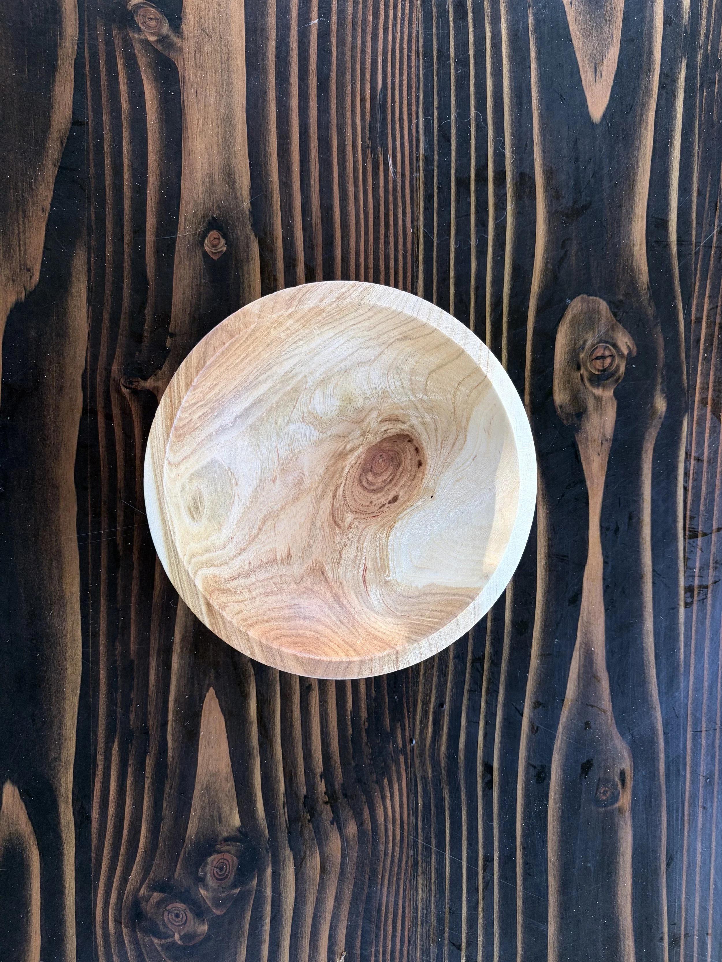 A round wooden plate with a natural wood grain pattern placed on a dark wood table with a striped pattern and visible knots.