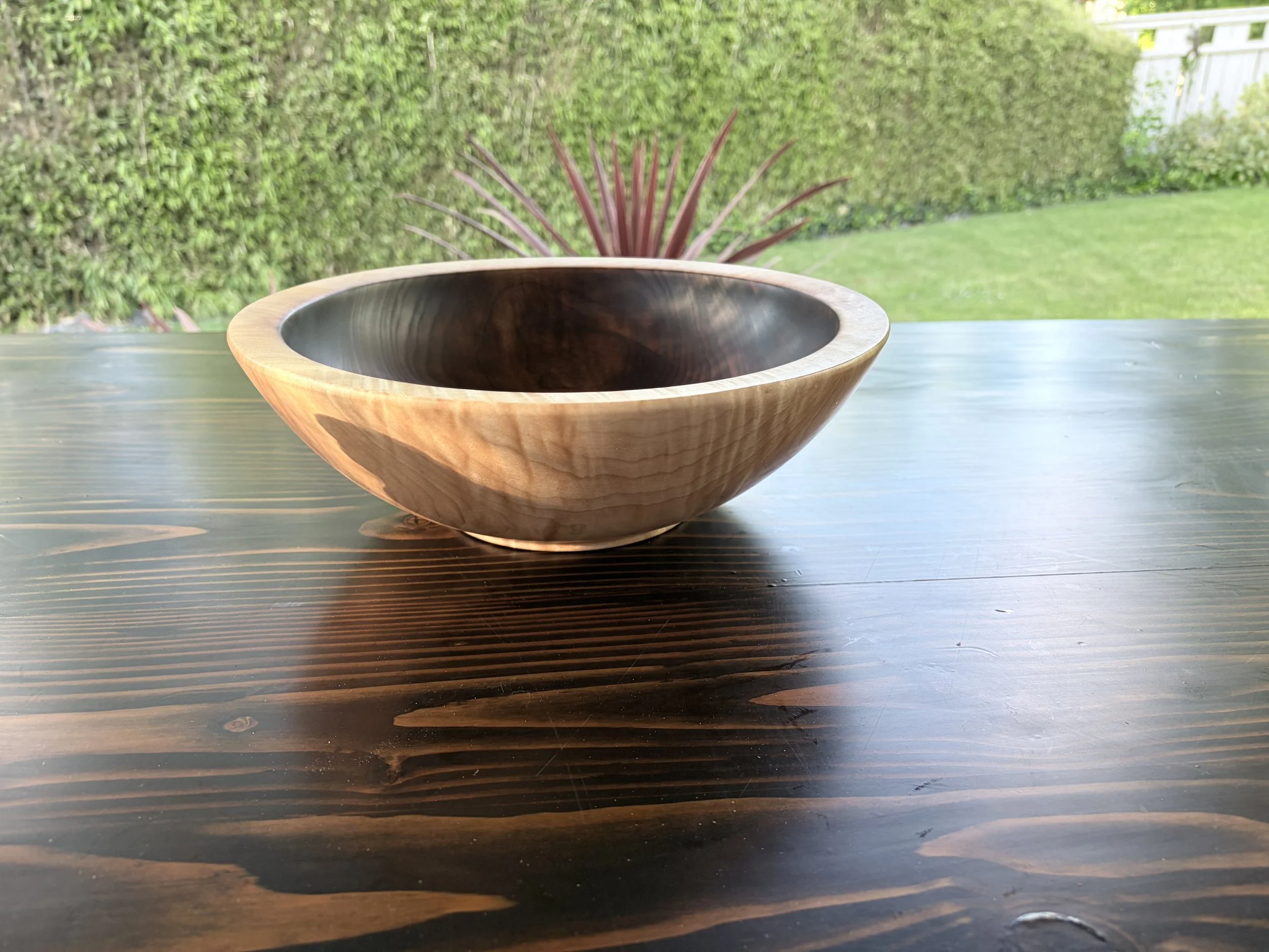 A wooden bowl with a dark interior sits on a wooden table outside, with a green hedge and out-of-focus plants in the background.
