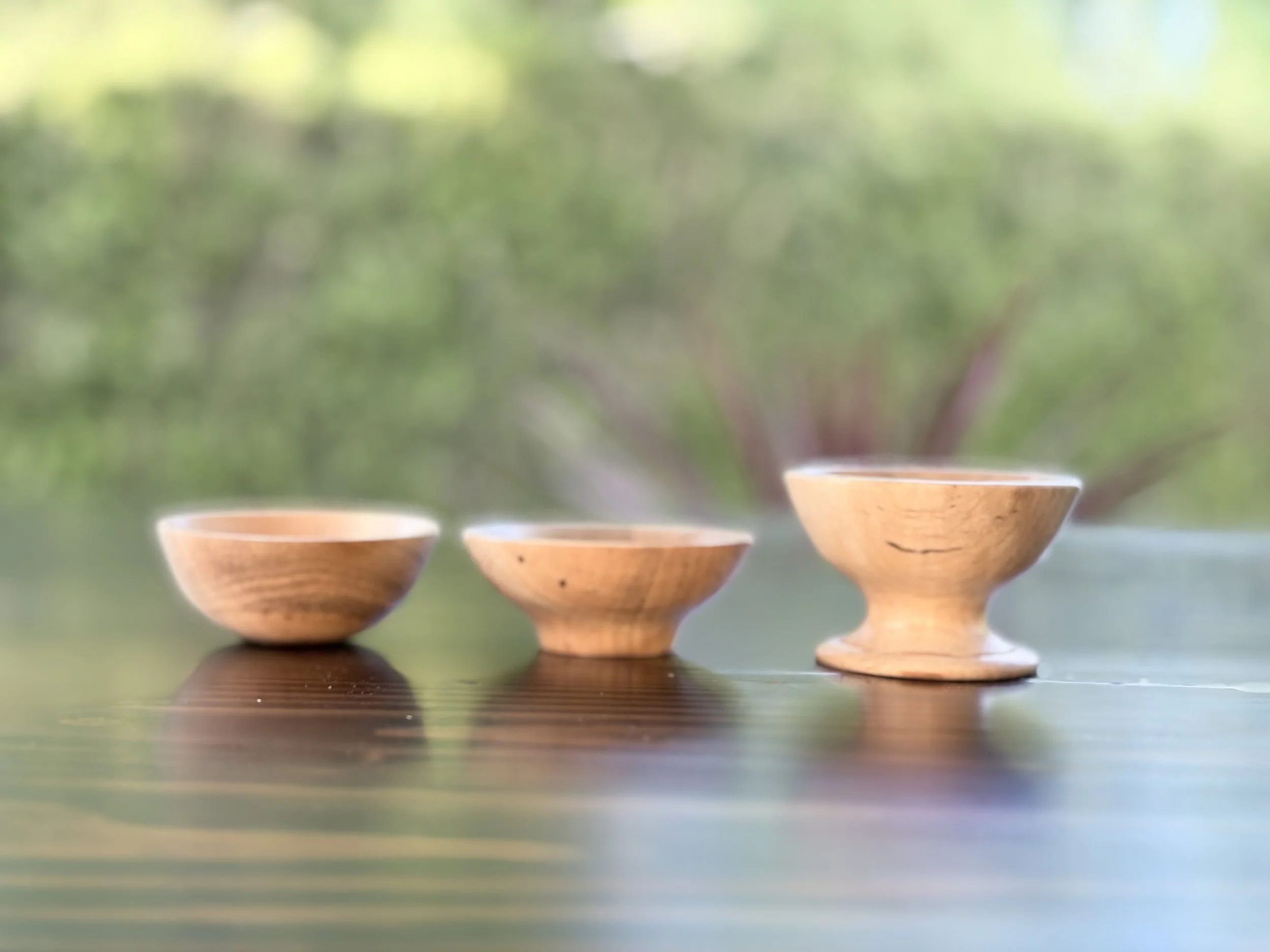 Three wooden bowls filled with a hot beverage, possibly tea, are arranged on a table outdoors with a blurred green background.