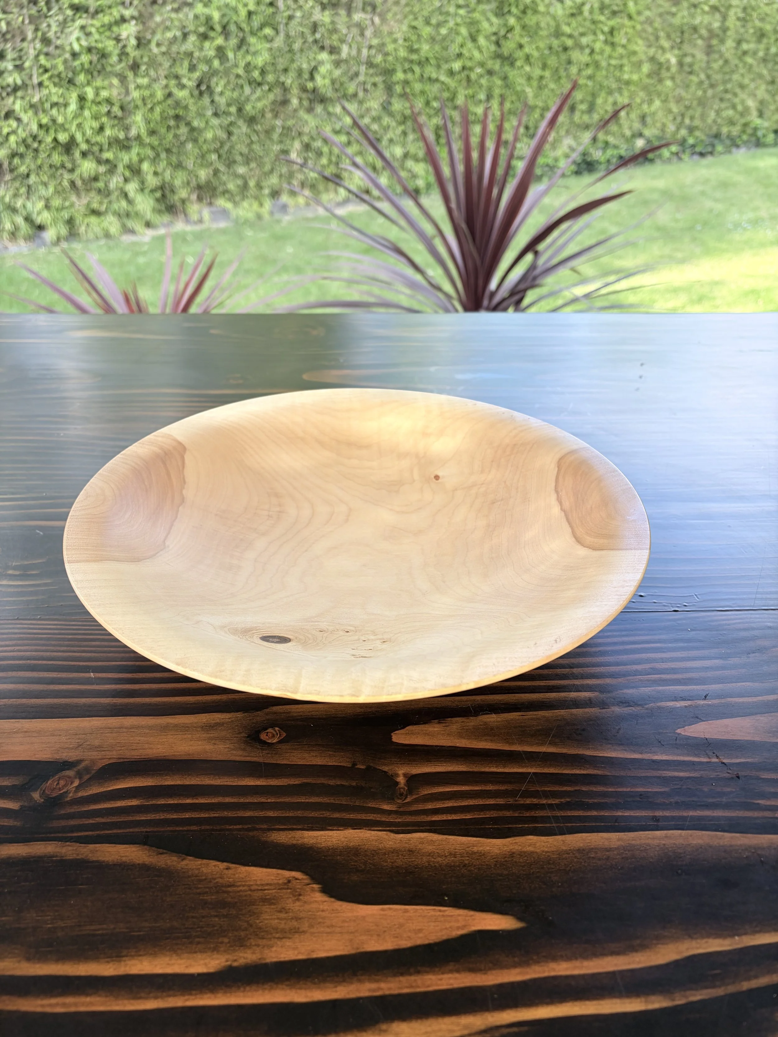 A round wooden bowl on a dark wooden table with an outdoor garden background of green bushes and plants.