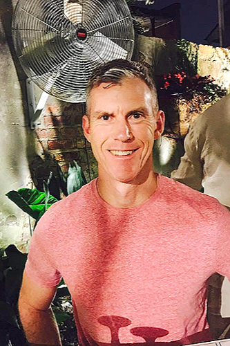 A man with short brown hair and a pink t-shirt smiling at the camera, standing outdoors near a large fan and some greenery.