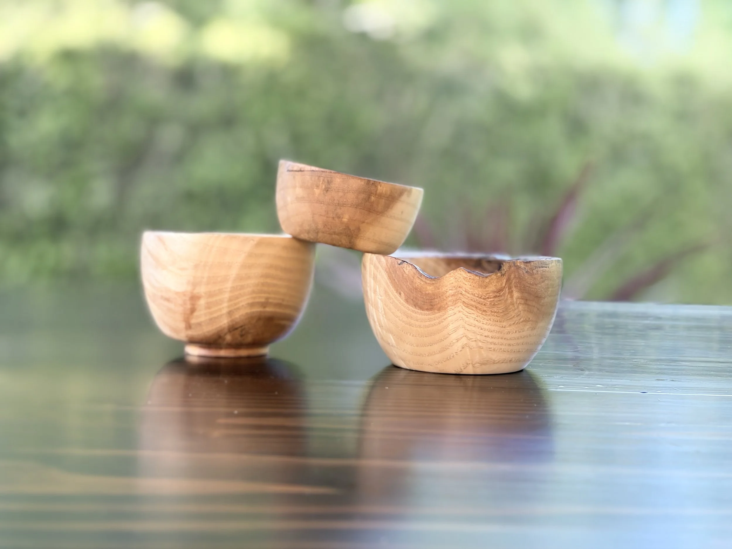 Close-up of three wooden bowls stacked on a dark wooden surface outdoors with green blurred background.