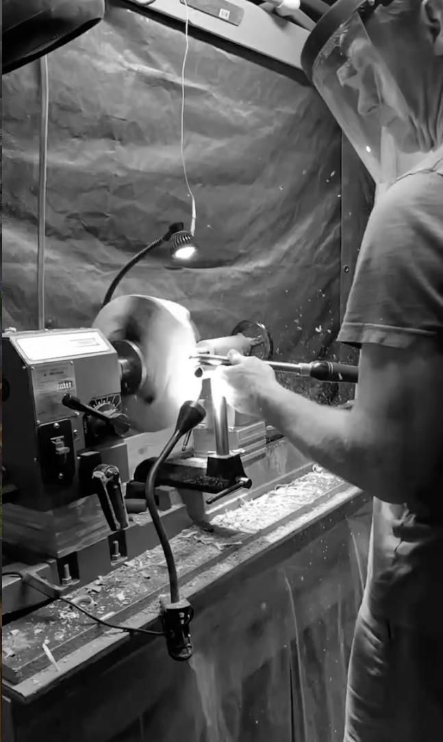 A person wearing a face shield and apron is operating a woodturning lathe, shaping a piece of wood.