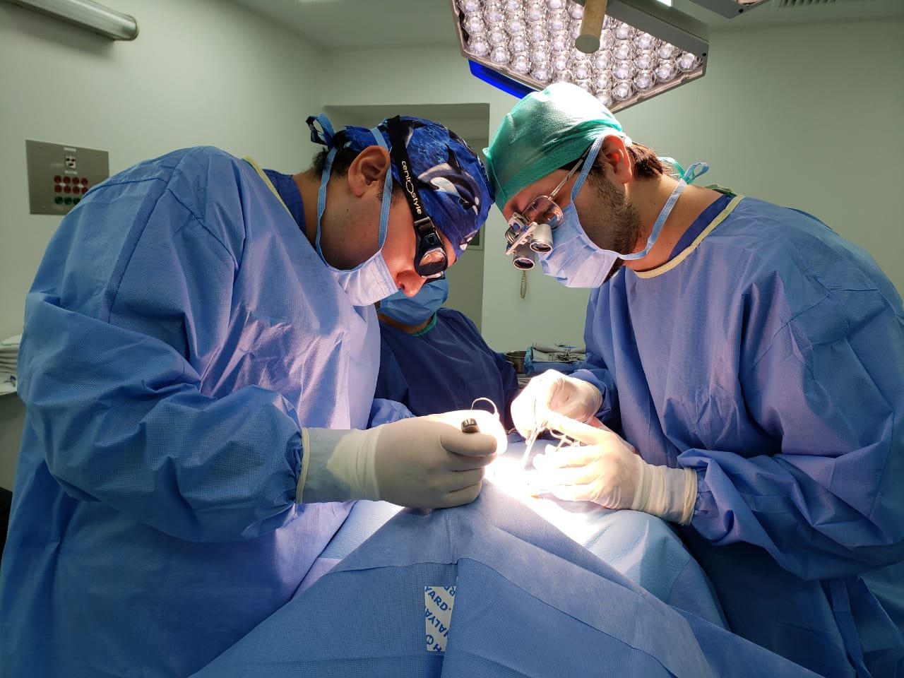Dr. Carlos Gustavo Rivera Robledo y su estudiante de subespecialidad en un quirófano realizando una operación de tiroides, usando batas, guantes, mascarillas y lentes de protección, con luz quirúrgica sobre ellos.
