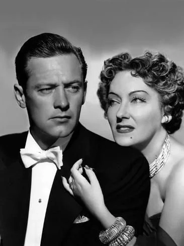 William Holden and Gloria Swanson in a publicity portrait for Sunset Boulevard (1950); Holden in a white bow tie and dinner jacket, Swanson behind him in a dark gown with layered pearls and a beaded bracelet, her hand resting on his chest.