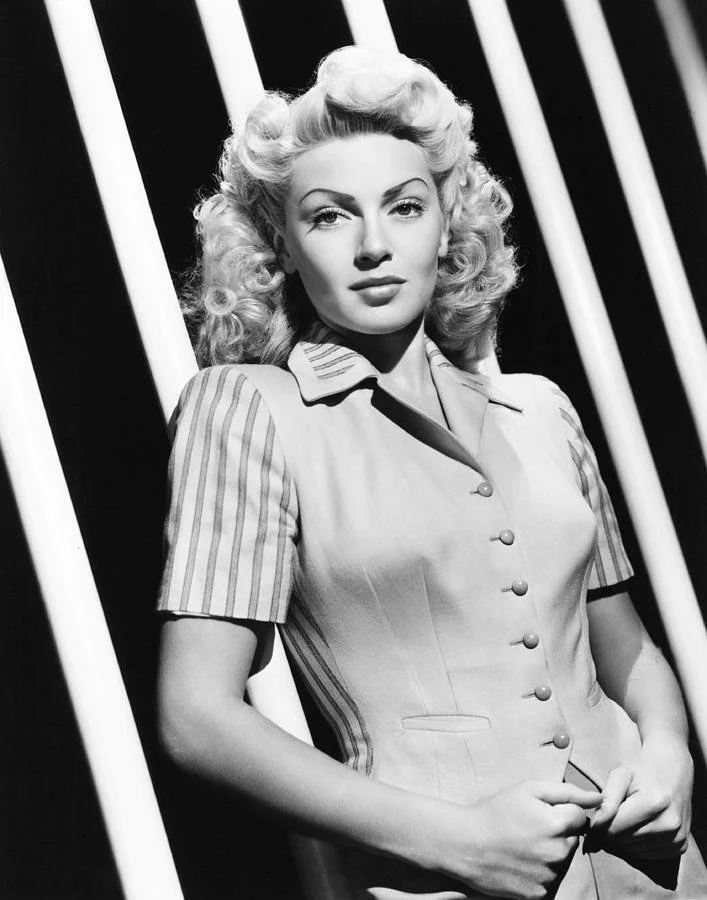 Lana Turner in a 1940s studio publicity portrait; platinum-blonde hair styled in curls, wearing a fitted button-front jacket with striped collar and sleeves, posed against a diagonal striped light-and-shadow backdrop.