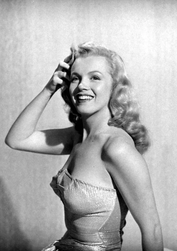 Black and white photo of a smiling woman with wavy blonde hair, wearing a strapless dress, touching her hair with one hand.