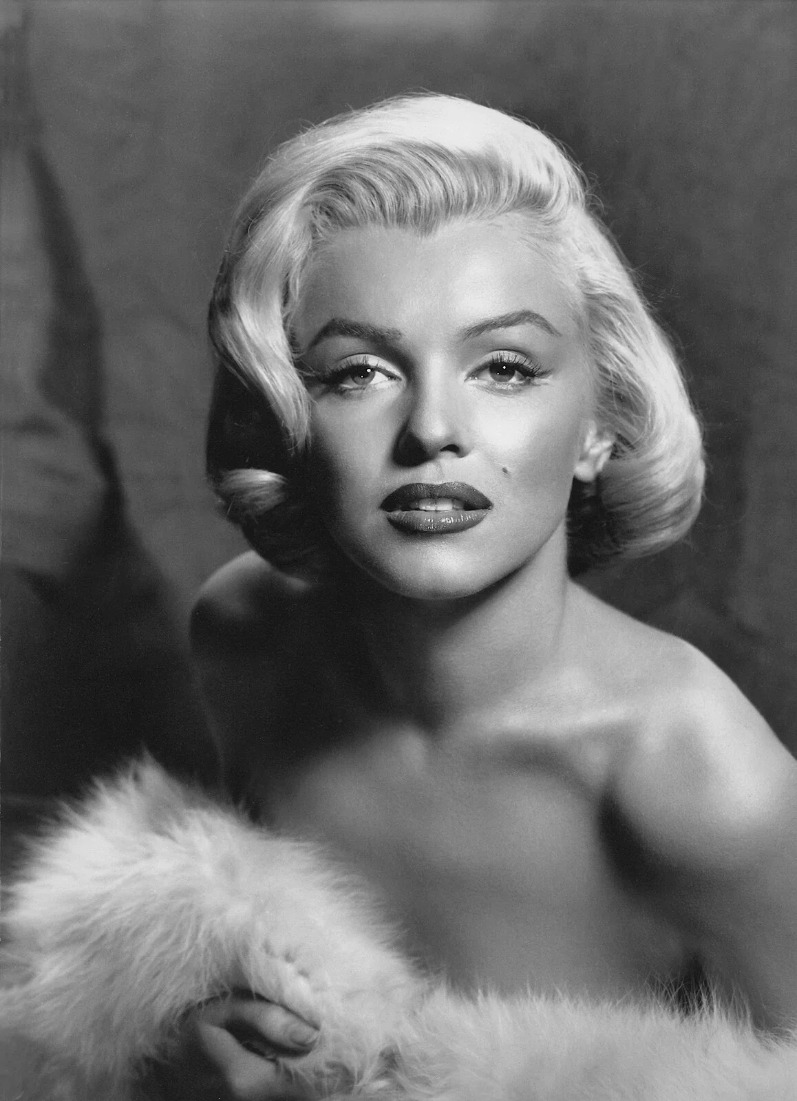 Black and white portrait of a woman with styled blonde hair, wearing lipstick, looking directly at the camera with a confident expression, and dressed in a fur garment.